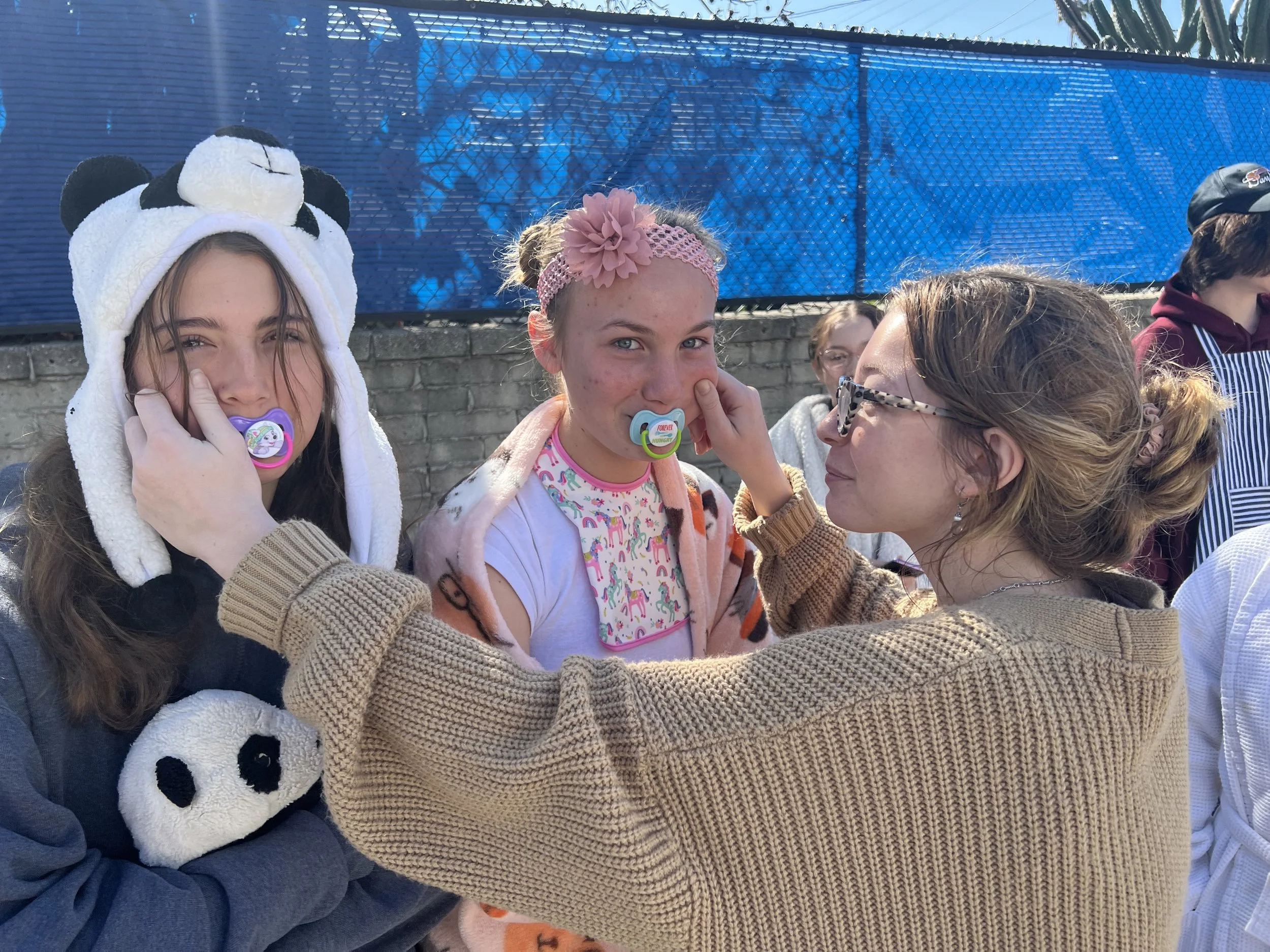 Group of children and an adult outdoors, with two girls wearing cartoon character-themed pacifiers and a girl with a pink headband, embracing and smiling.