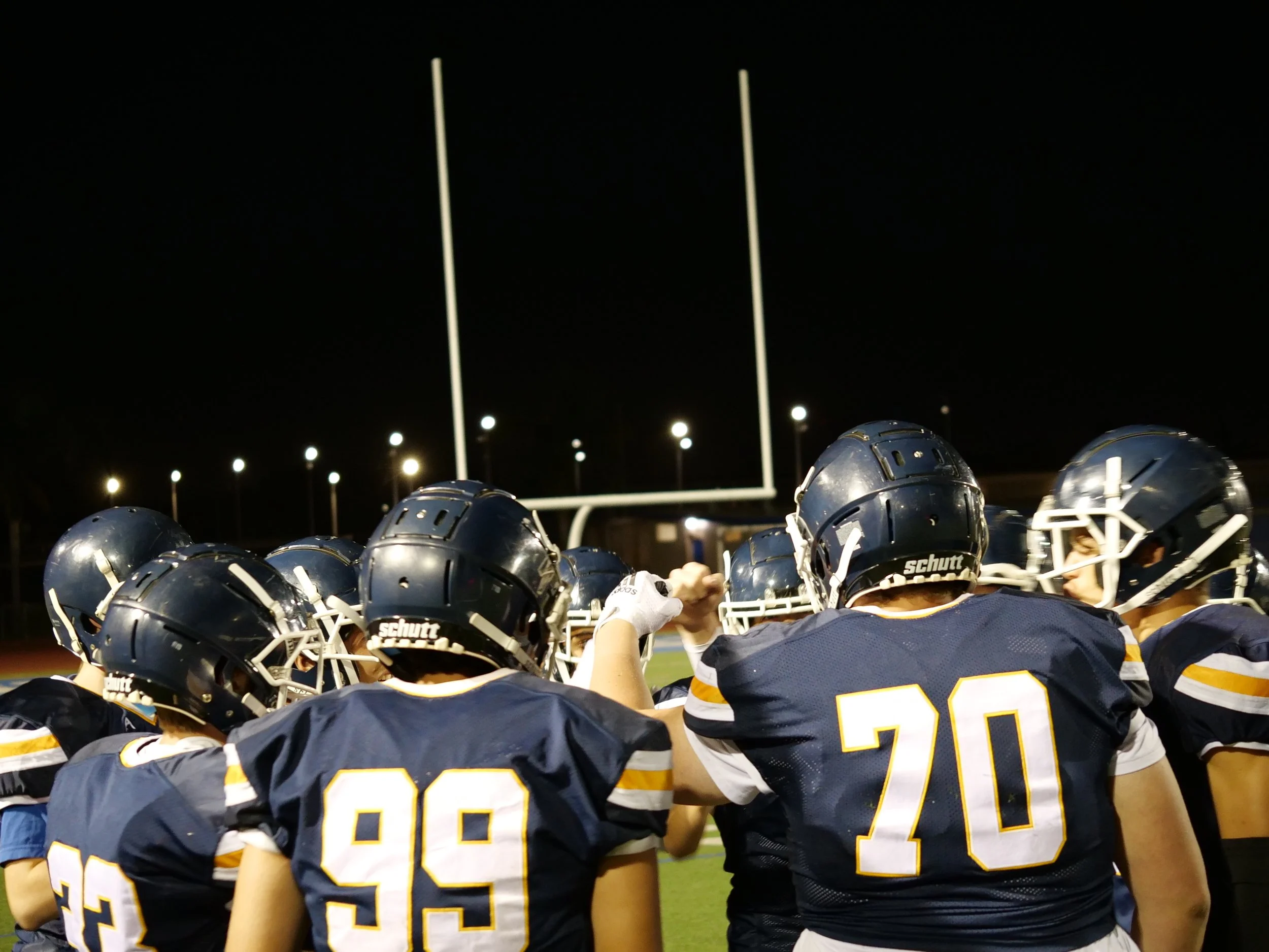 A group of football players wearing navy blue and yellow uniforms and helmets, huddled together on the field at night, with goalposts and bright stadium lights in the background.