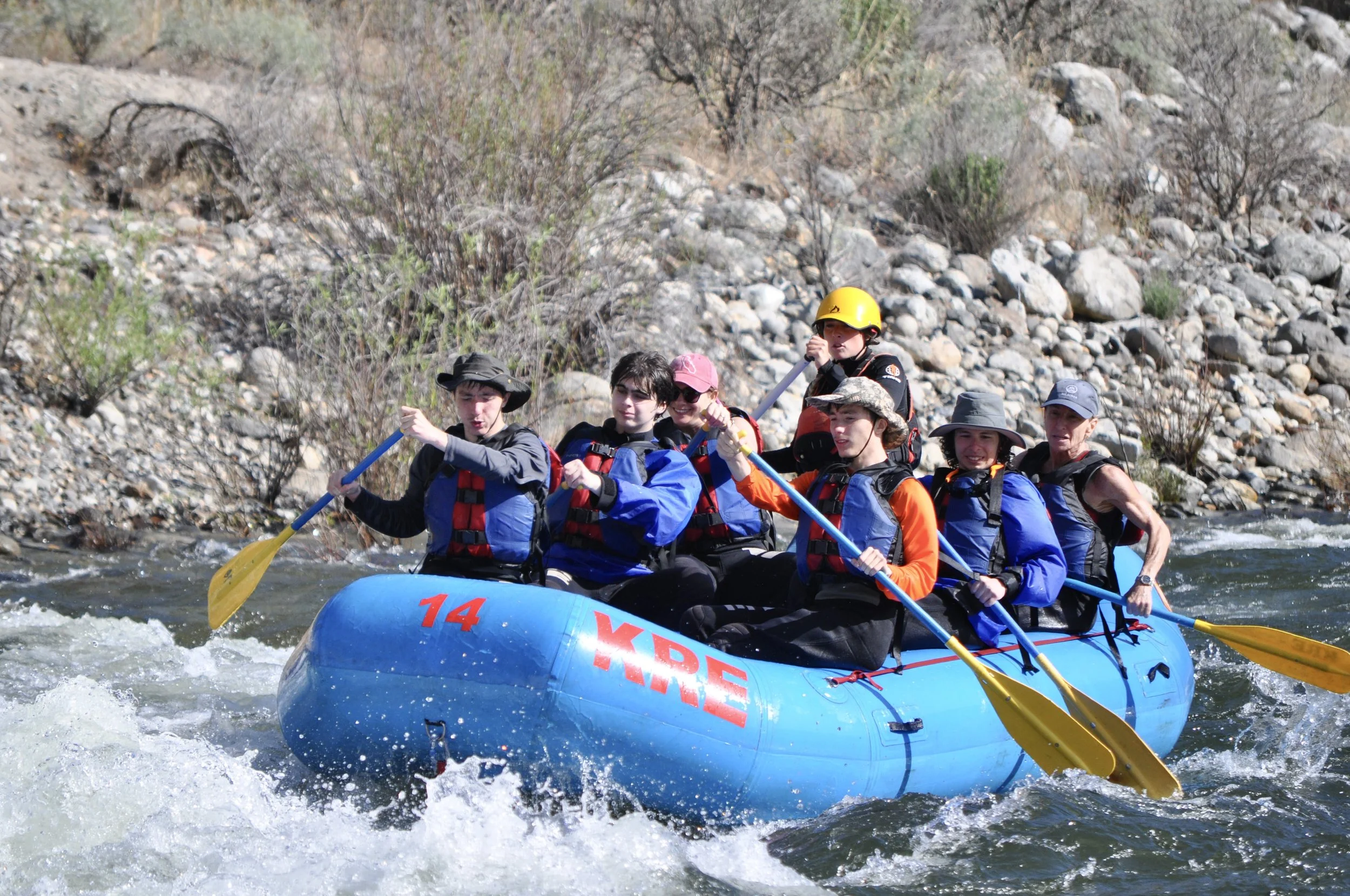 Group of people white water rafting in a rocky river, wearing life jackets and helmets.