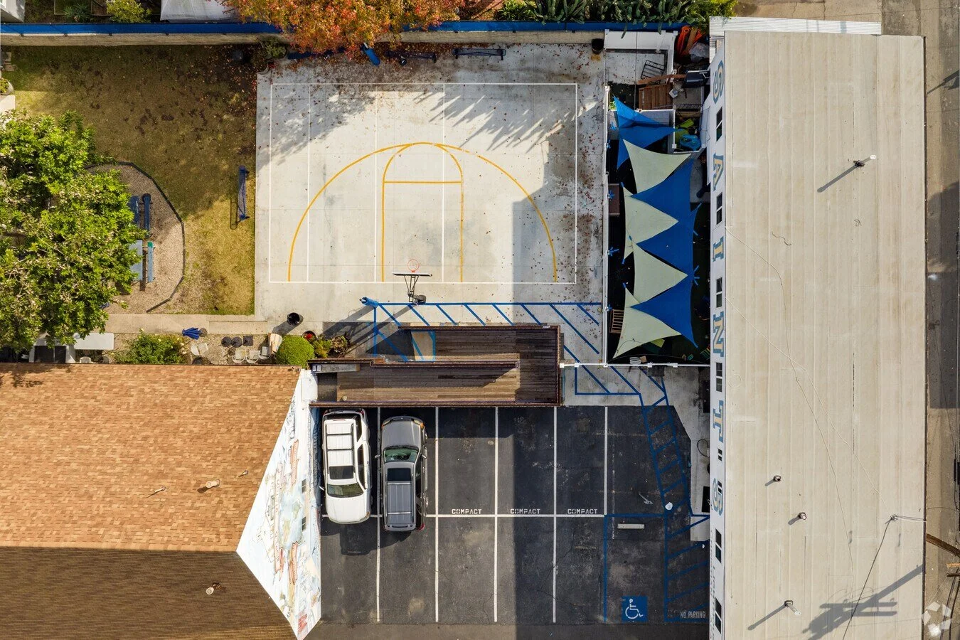 An aerial view of a small urban area showing a basketball court, a parking lot with two cars, a building with a mural, and a section with blue and green tents.