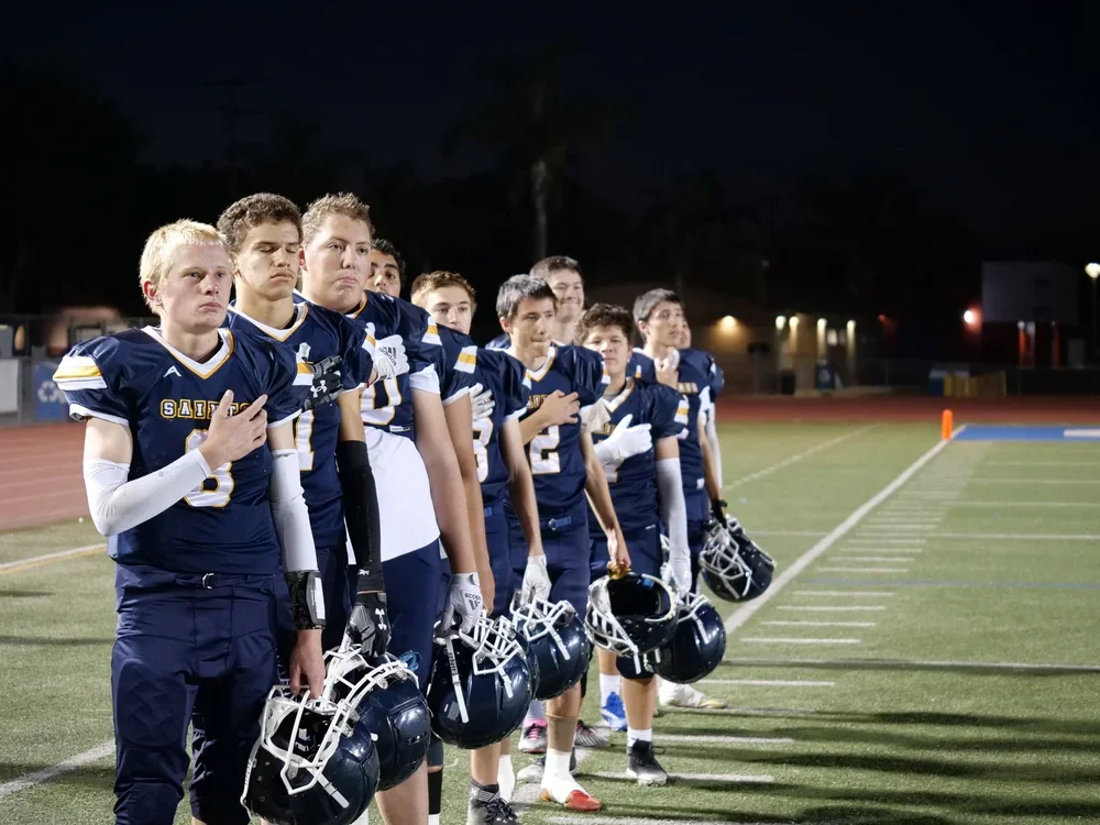 A group of boys in football uniforms standing on a football field during a nighttime game or event, facing forward with some holding helmets.