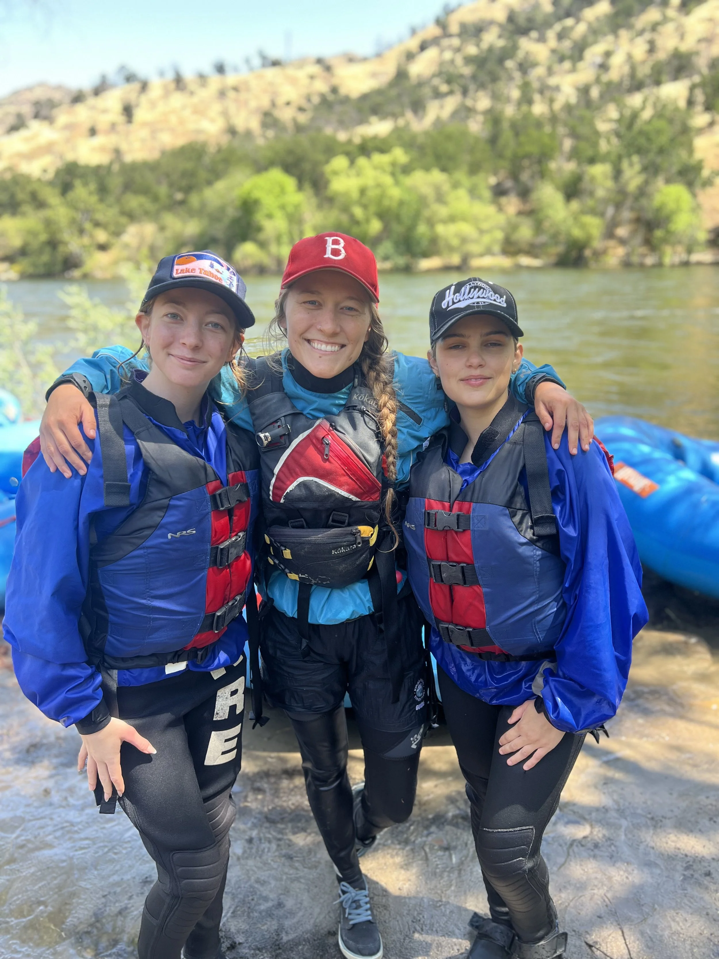 Three women in outdoor gear standing together near a river, smiling, with green trees and hills in the background.