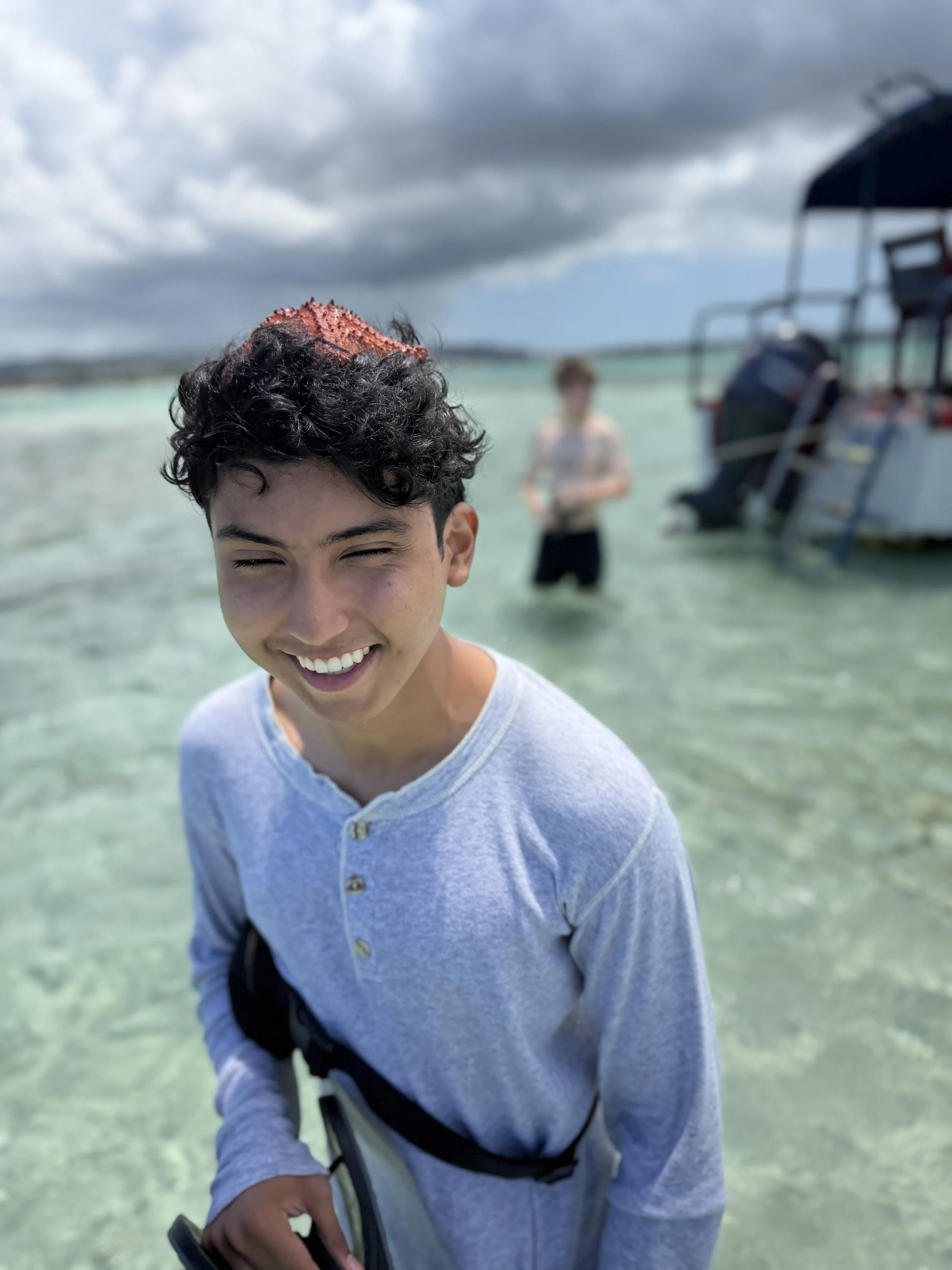A smiling young man with curly dark hair standing in shallow clear water at the beach, with a younger person in the background near a boat and cloudy skies above.