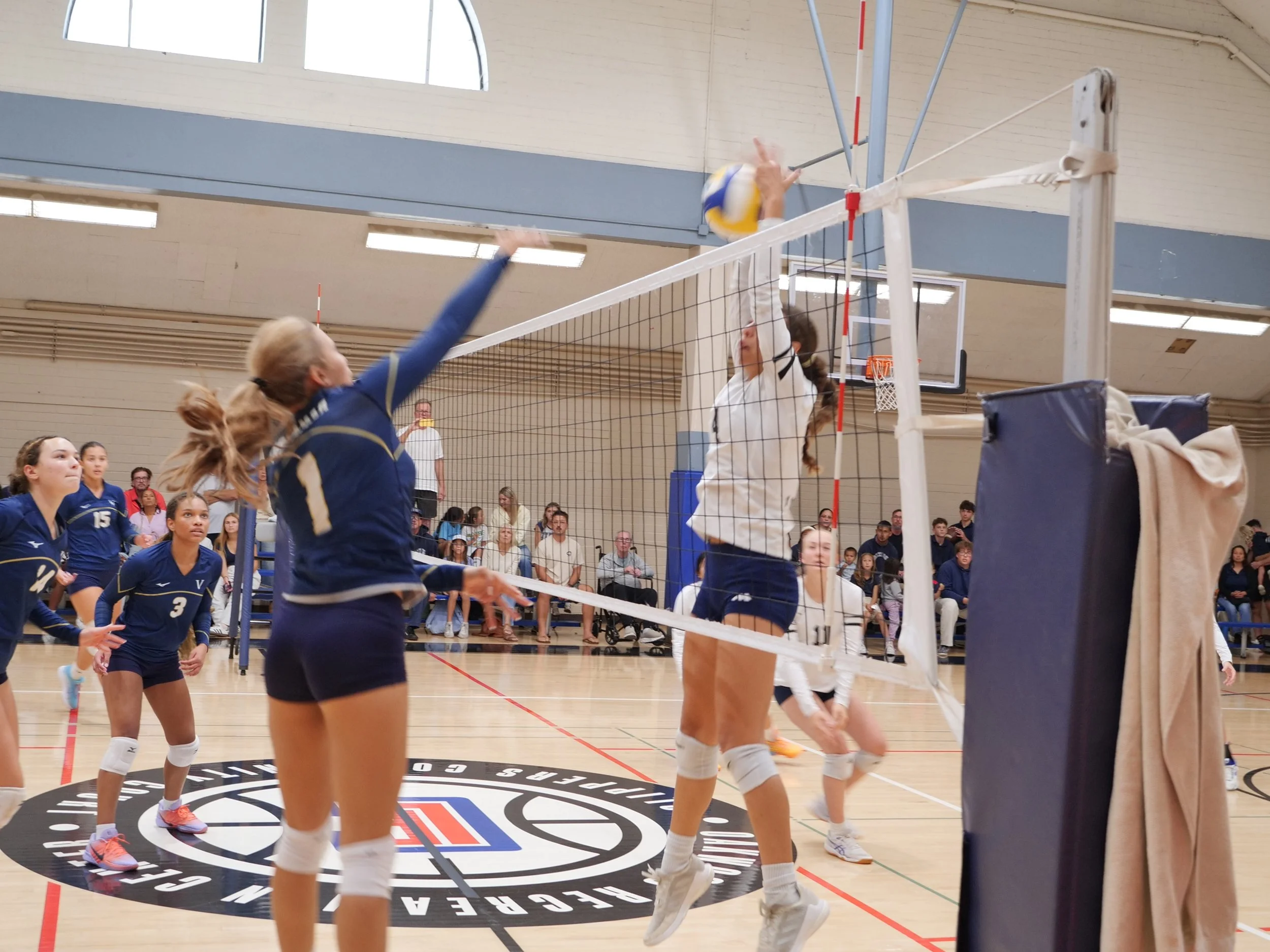 Indoor volleyball game with players jumping to hit the ball over the net, spectators watching in the background.