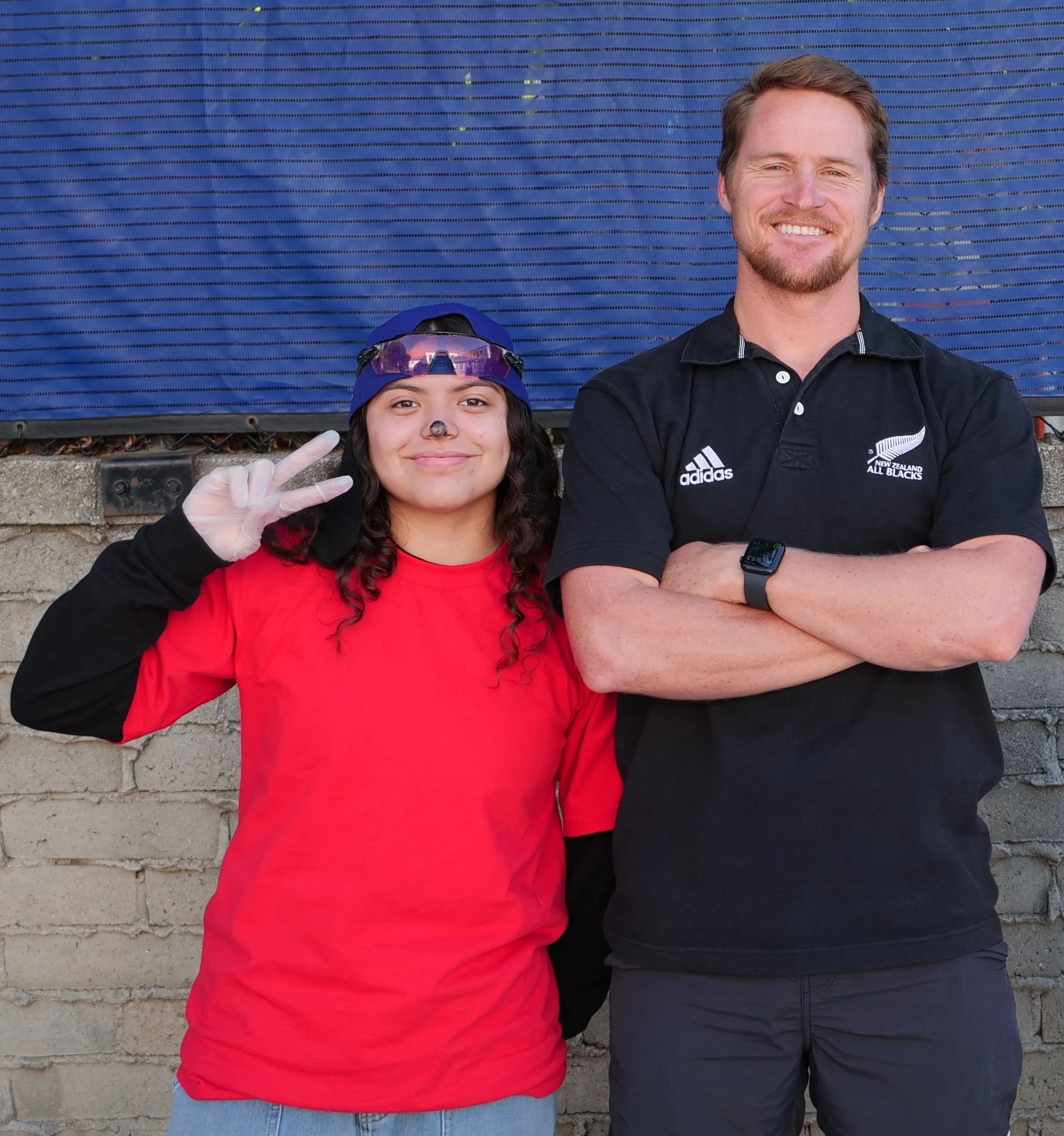 Two people standing next to a brick wall, smiling, with one person wearing a red shirt and gray jeans and the other wearing a black polo shirt with white logo and black pants, one holds up a peace sign, and the background features a large blue surfac