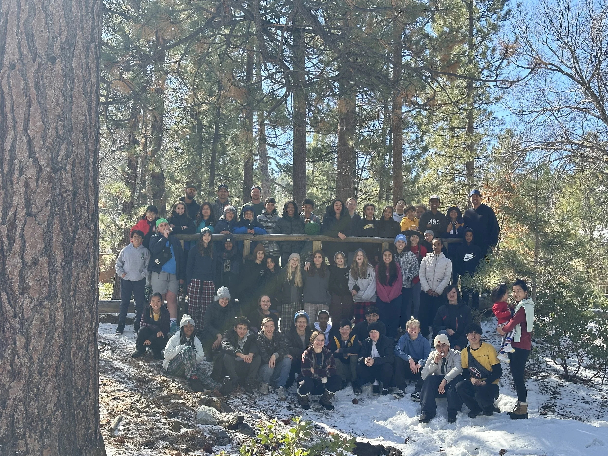 A large group of people, mostly children and teenagers, posing together outdoors in a forested area with tall pine trees and snow on the ground. The group is smiling and some are sitting while others are standing, with a few adults present.
