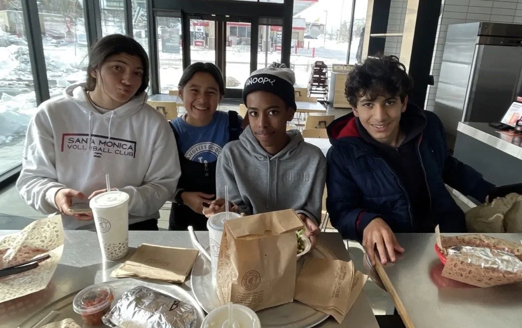 Four teenagers sitting at a table in a restaurant, with pizza, drinks, and food containers in front of them, near large windows showing a snowy outdoor scene.