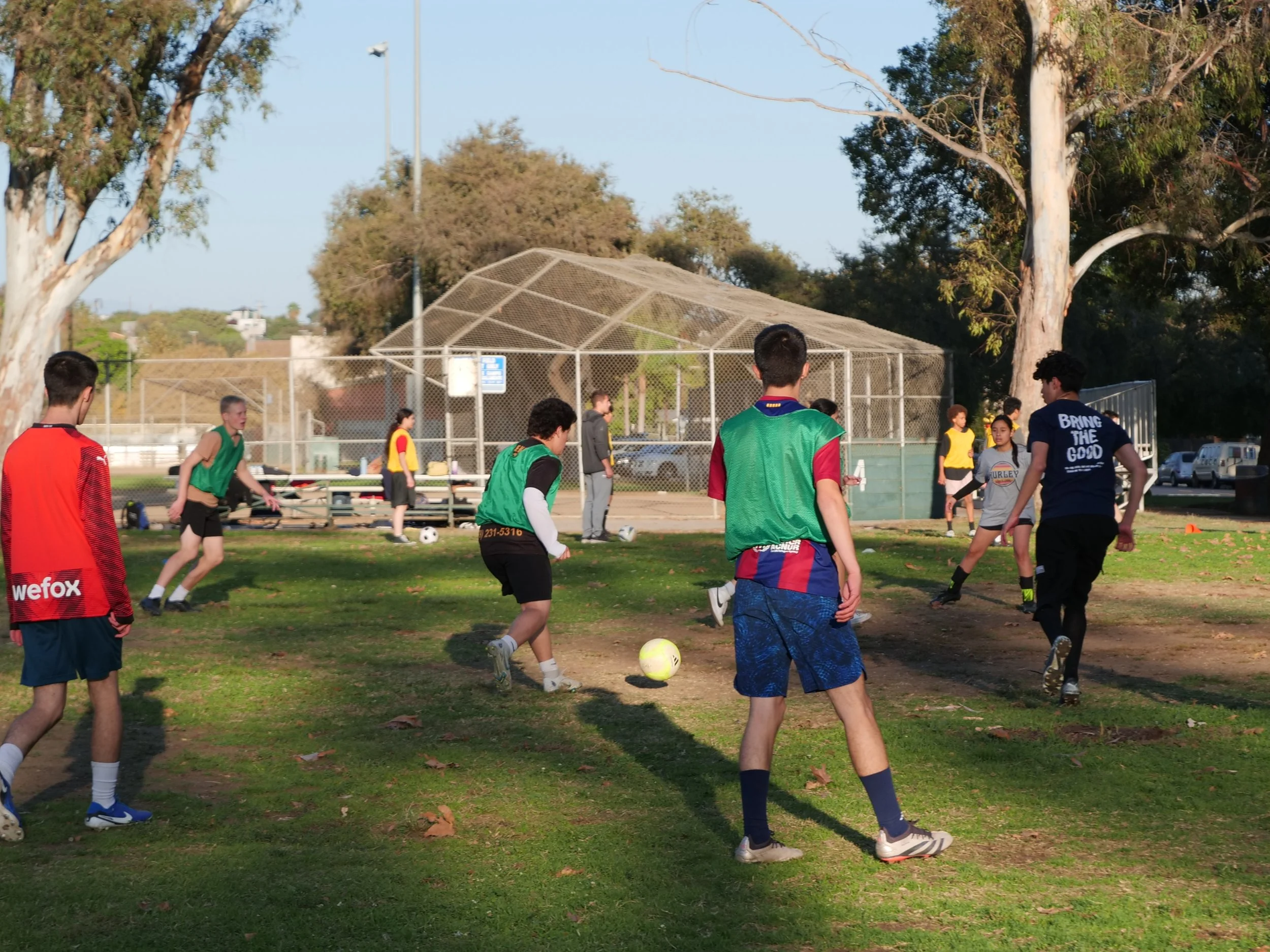 Group of children playing soccer outdoors on a grassy field, surrounded by trees and a chain-link fence, with some adults nearby.