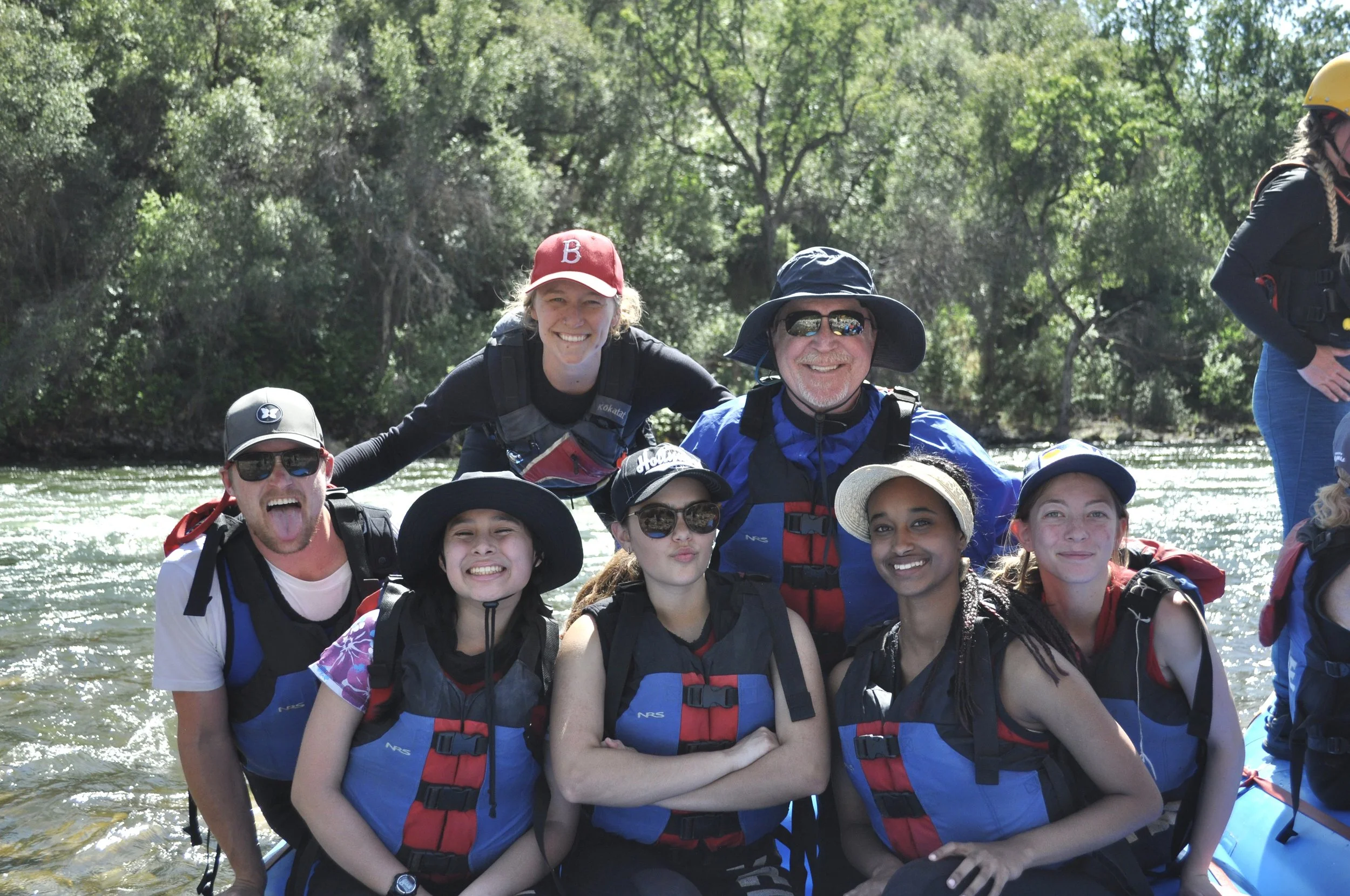 Group of smiling people in life jackets on a boat in a river surrounded by trees.