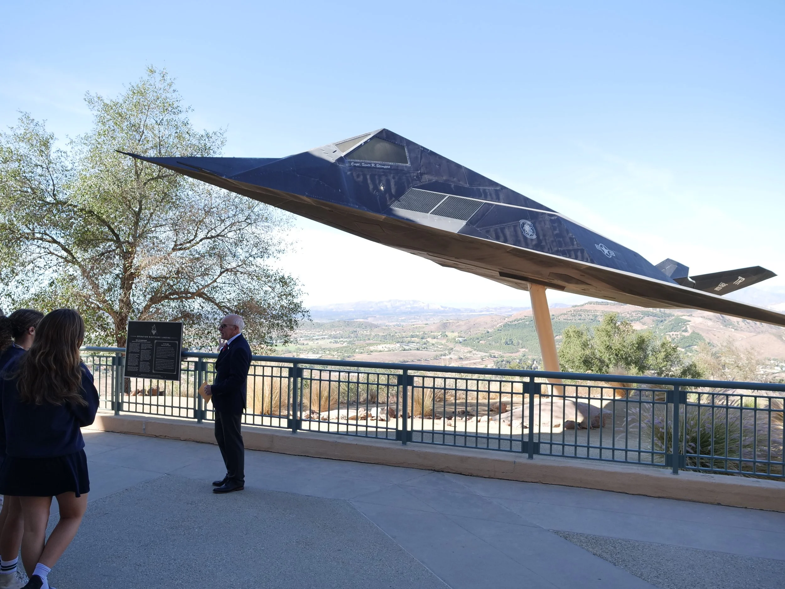 A sleek black aircraft displayed outdoors on a stand with a desert landscape in the background. Several people are observing the aircraft, with some engaged in conversation.