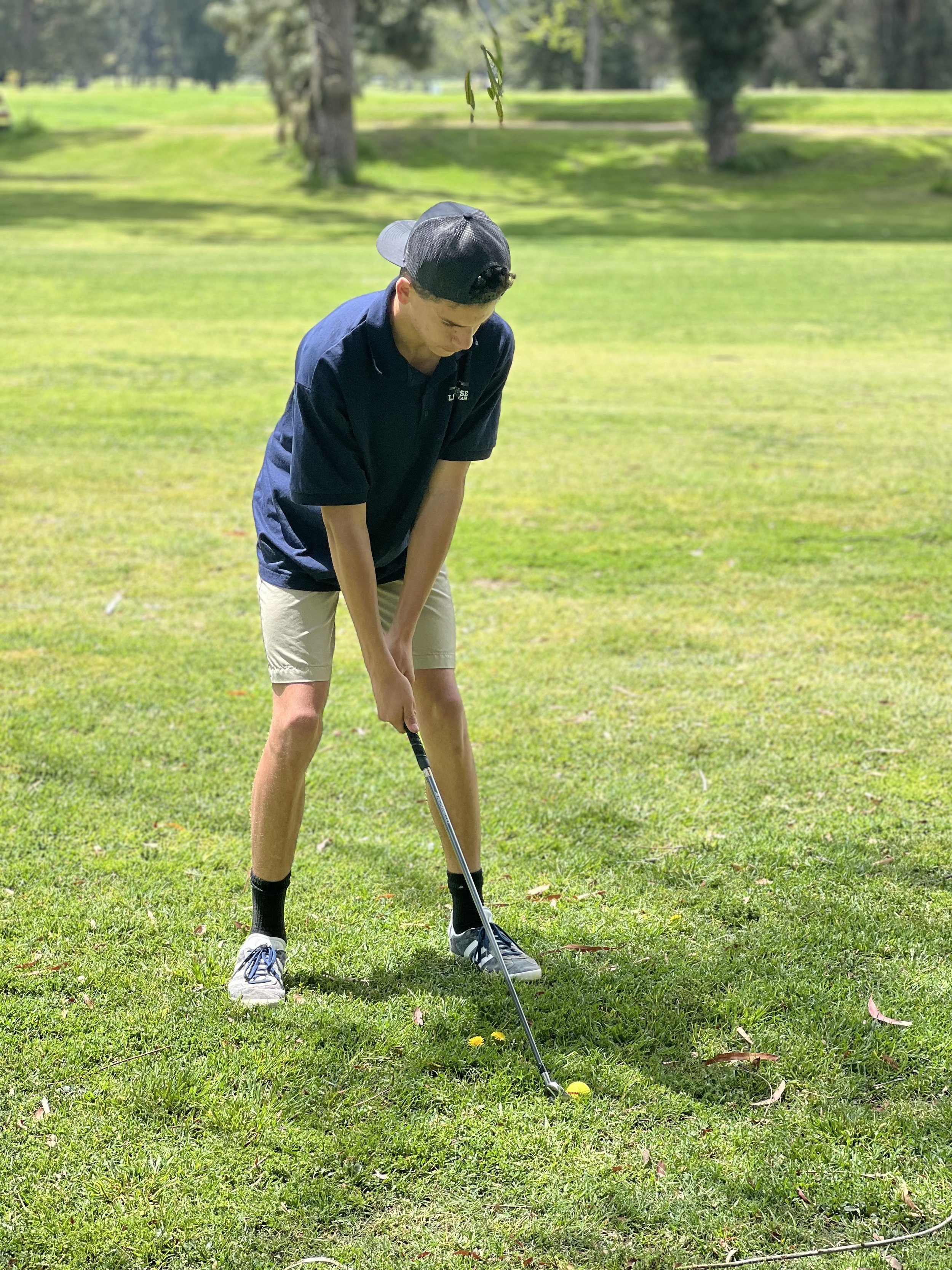 A young man playing golf on a grassy golf course, preparing to hit a golf ball with a club, wearing a black cap, navy shirt, khaki shorts, and sneakers.
