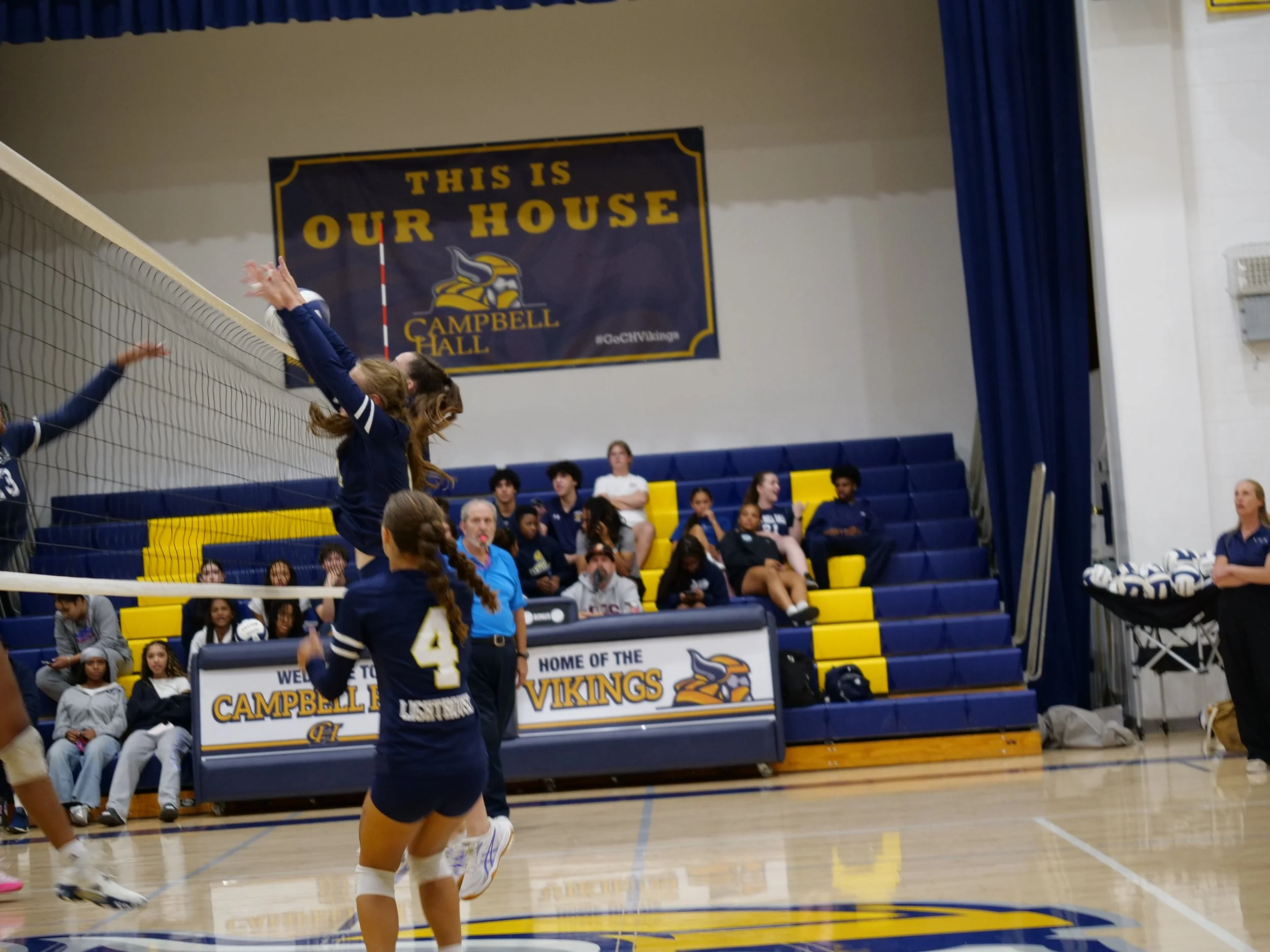 Indoor volleyball game with players jumping near the net, wearing blue uniforms, in a gymnasium. A sign reads 'This is Our House Campbell Hall' with the Vikings logo in the background, and spectators watching from bleachers.