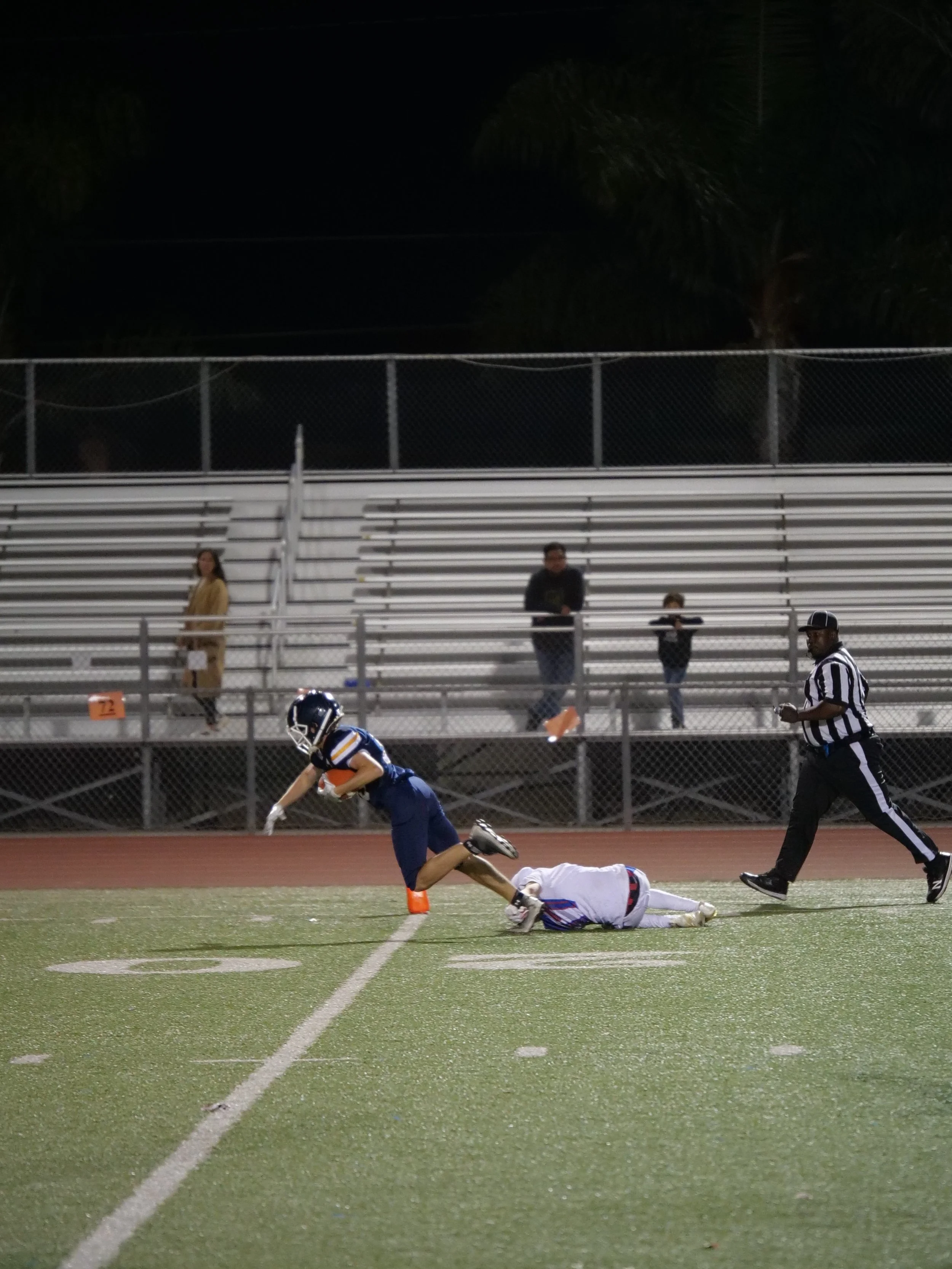 A football player in a blue uniform diving to catch the ball while a player in white lies on the field during a game at night, with officials and spectators in the background.