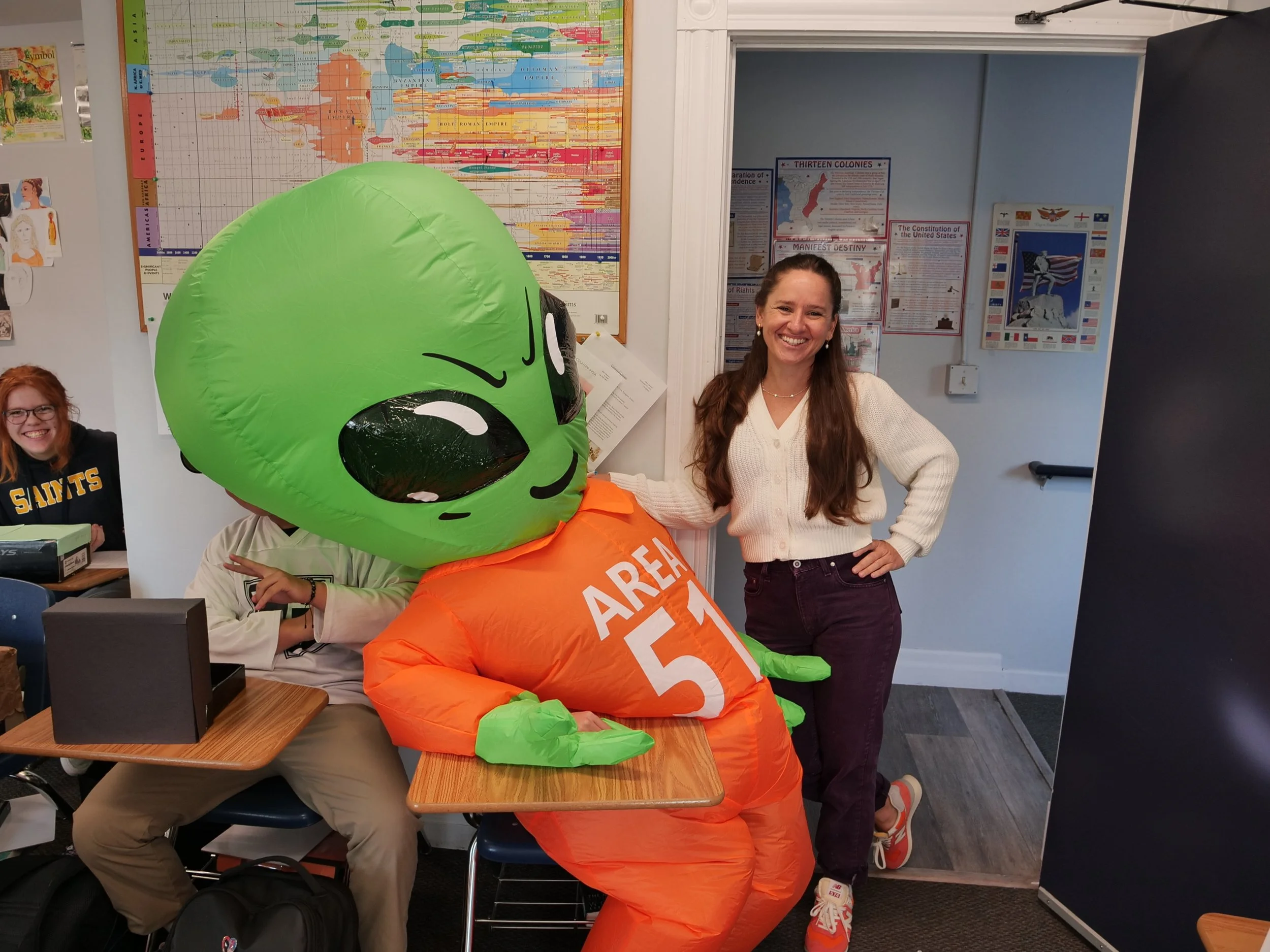 A woman smiling and standing next to a person in an alien mascot costume in a classroom. The mascot is wearing an orange outfit labeled 'AREA 51' and has a large green head. The classroom walls have posters, a world map, and a student sitting at a desk.