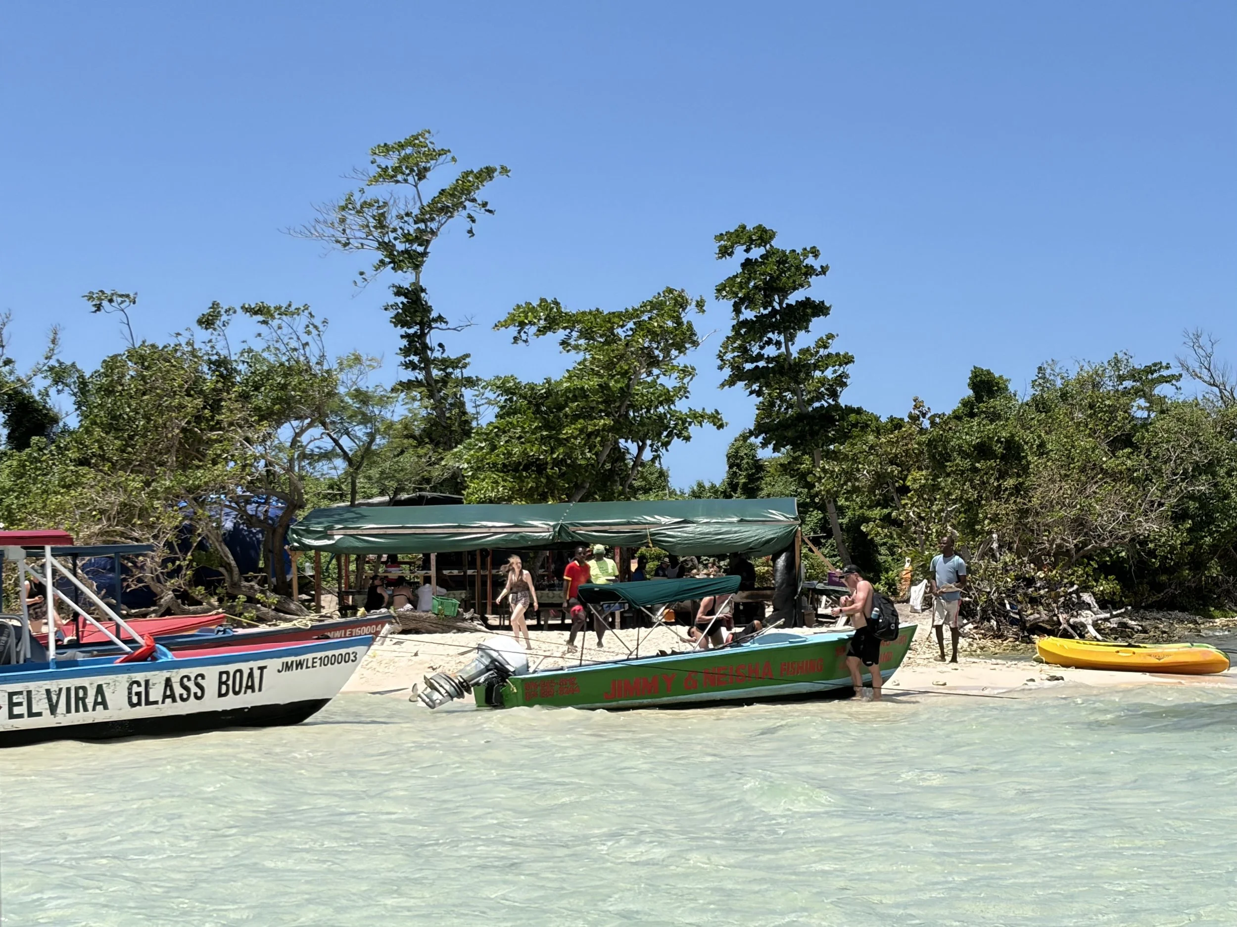 Fishermen's boats on a sandy beach near scattered trees, some boats have names and registration numbers, and a small group of people is visible near the boats under a canopy, with a bright blue sky above.