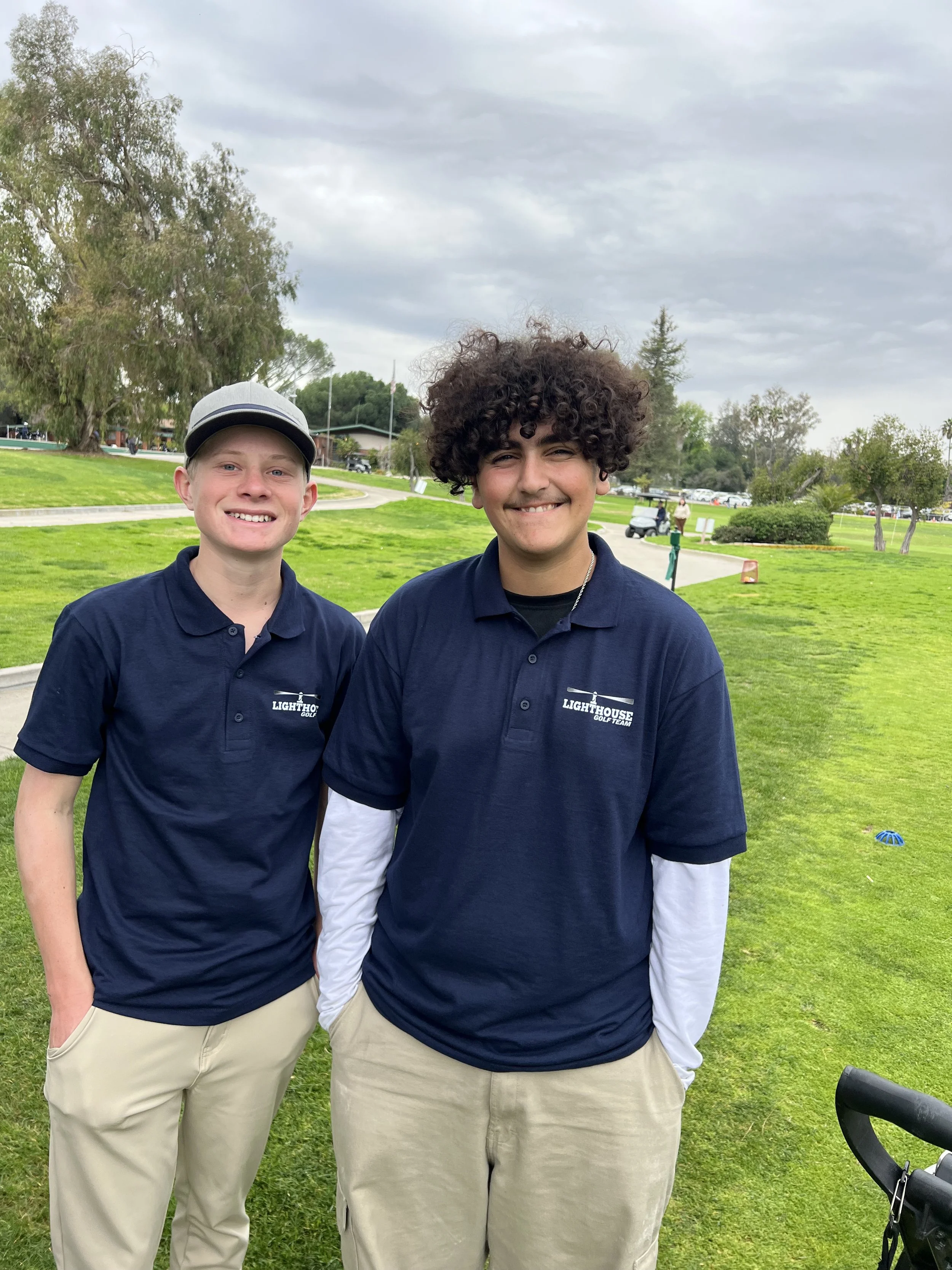 Two young men standing outdoors on a golf course, smiling. They are wearing navy blue polo shirts with 'Lighthouse GOLF TEAM' logo, beige pants, and the man on the left wears a gray hat. The background features trees, a golf cart path, and cloudy sky.