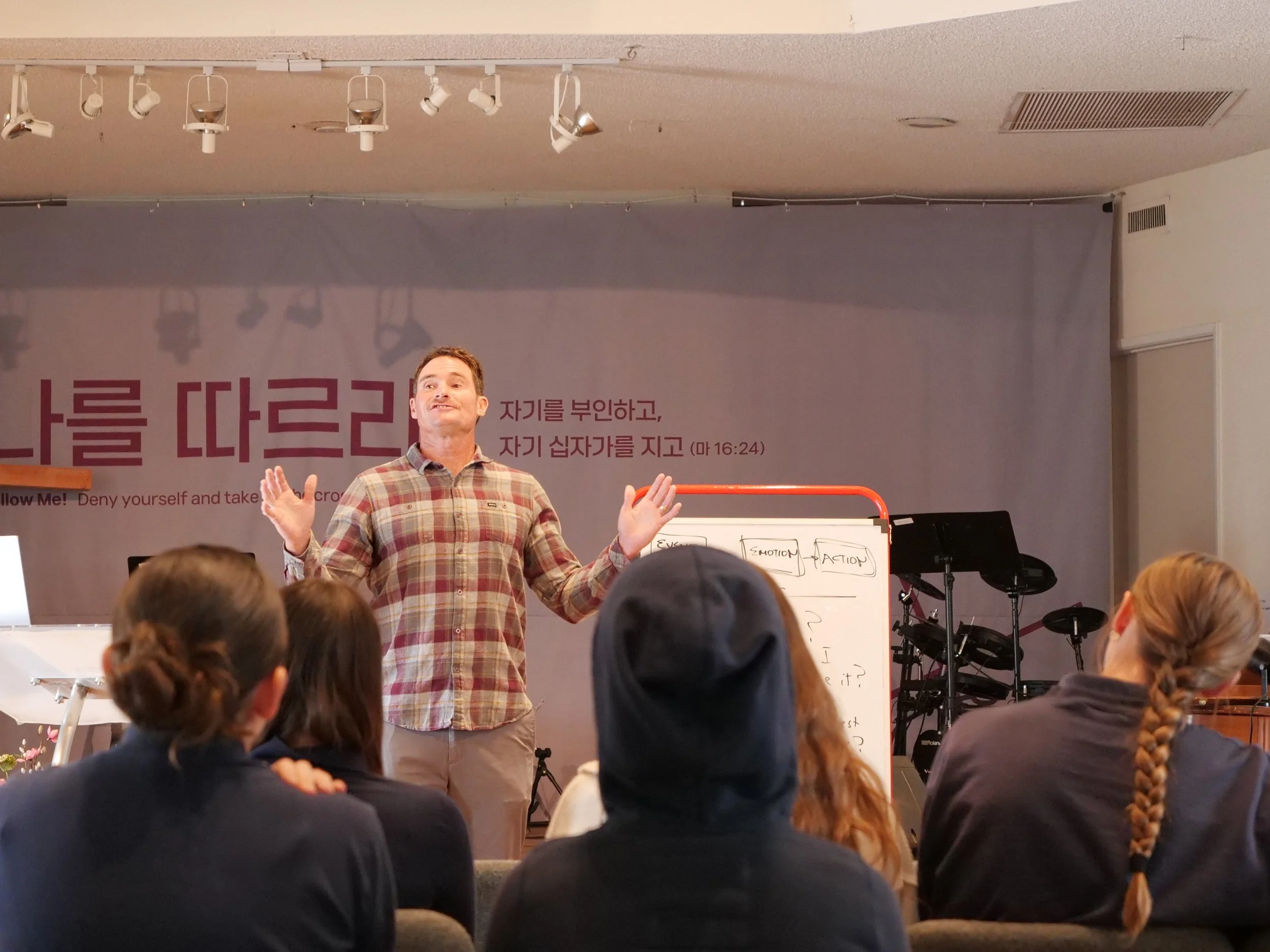 A man in a plaid shirt speaking to an audience in a classroom or seminar setting, with a purple presentation screen and a whiteboard nearby. Multiple audience members are visible from behind.