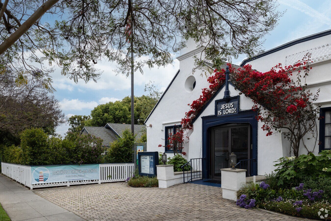 Exterior of Lighthouse Church and School with white building, black accents, red flowering vines, greenery, and a sign that reads "JESUS IS LORD!"