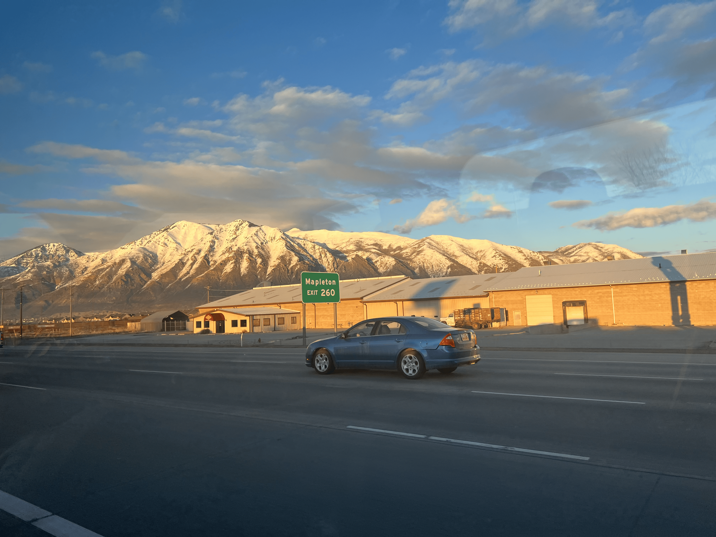 A scene of snow-capped mountains under a sky with scattered clouds, viewed from a parking lot with several buildings and a gray car in the foreground. There is also a green road sign that reads 'Mapleton EXIT 260'.