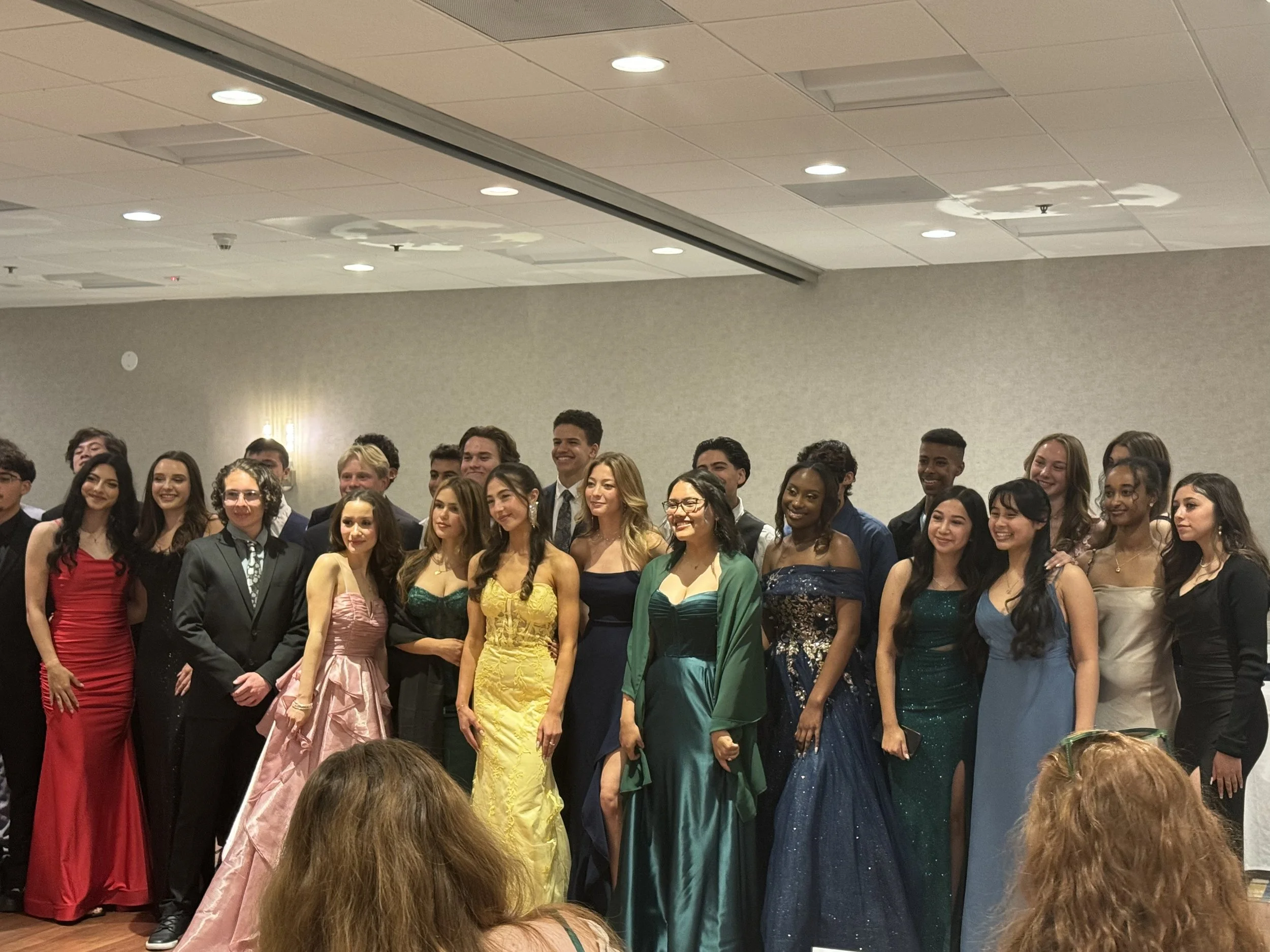 Group of young adults dressed in formal and evening wear, smiling and posing together at an event, standing in front of a beige wall and ceiling with overhead lights.