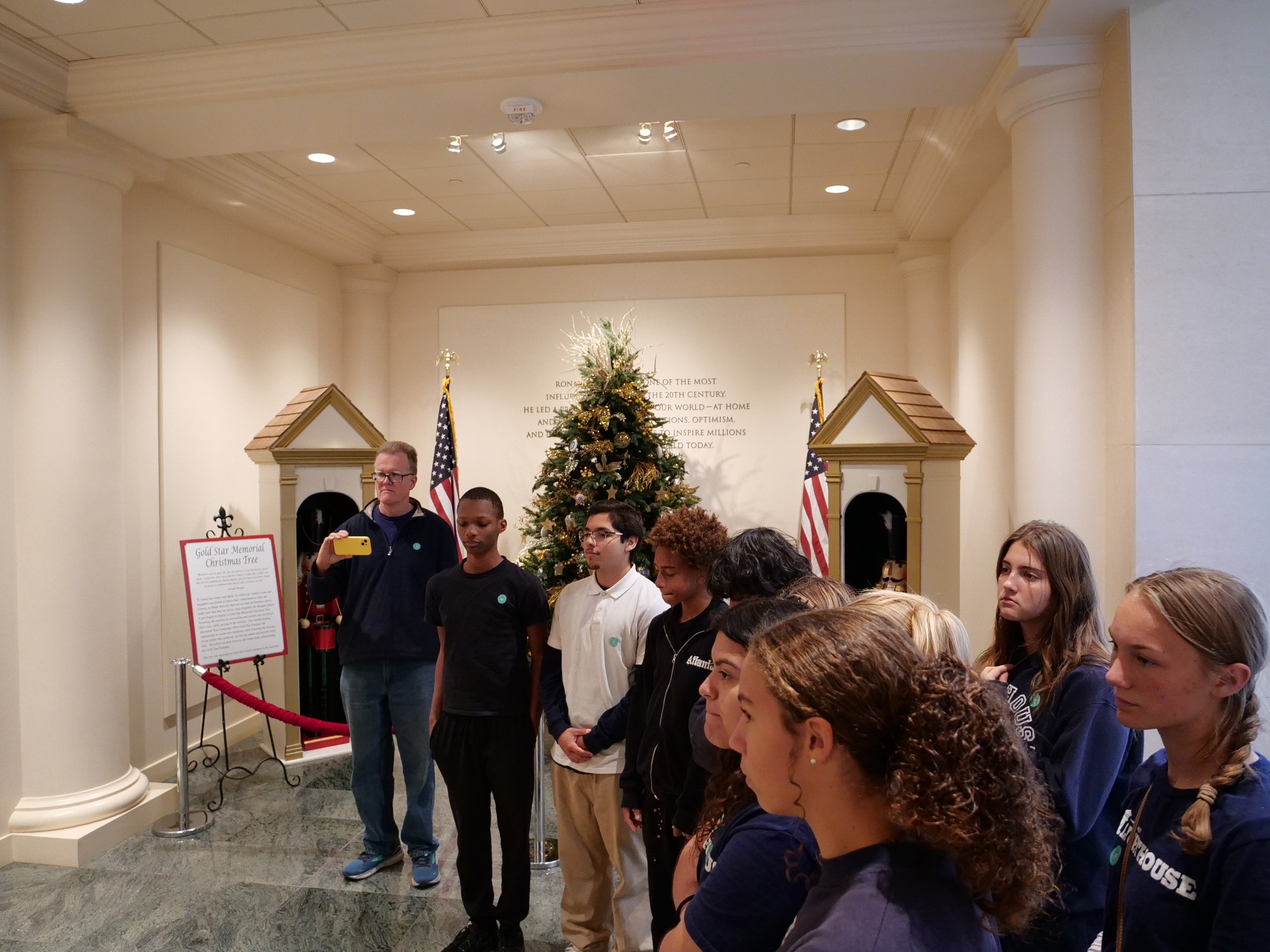 A group of people, including children and adults, standing in a line in front of a Christmas tree in a room with beige walls and columns. There are American flags and a sign about a Christmas tree nearby.
