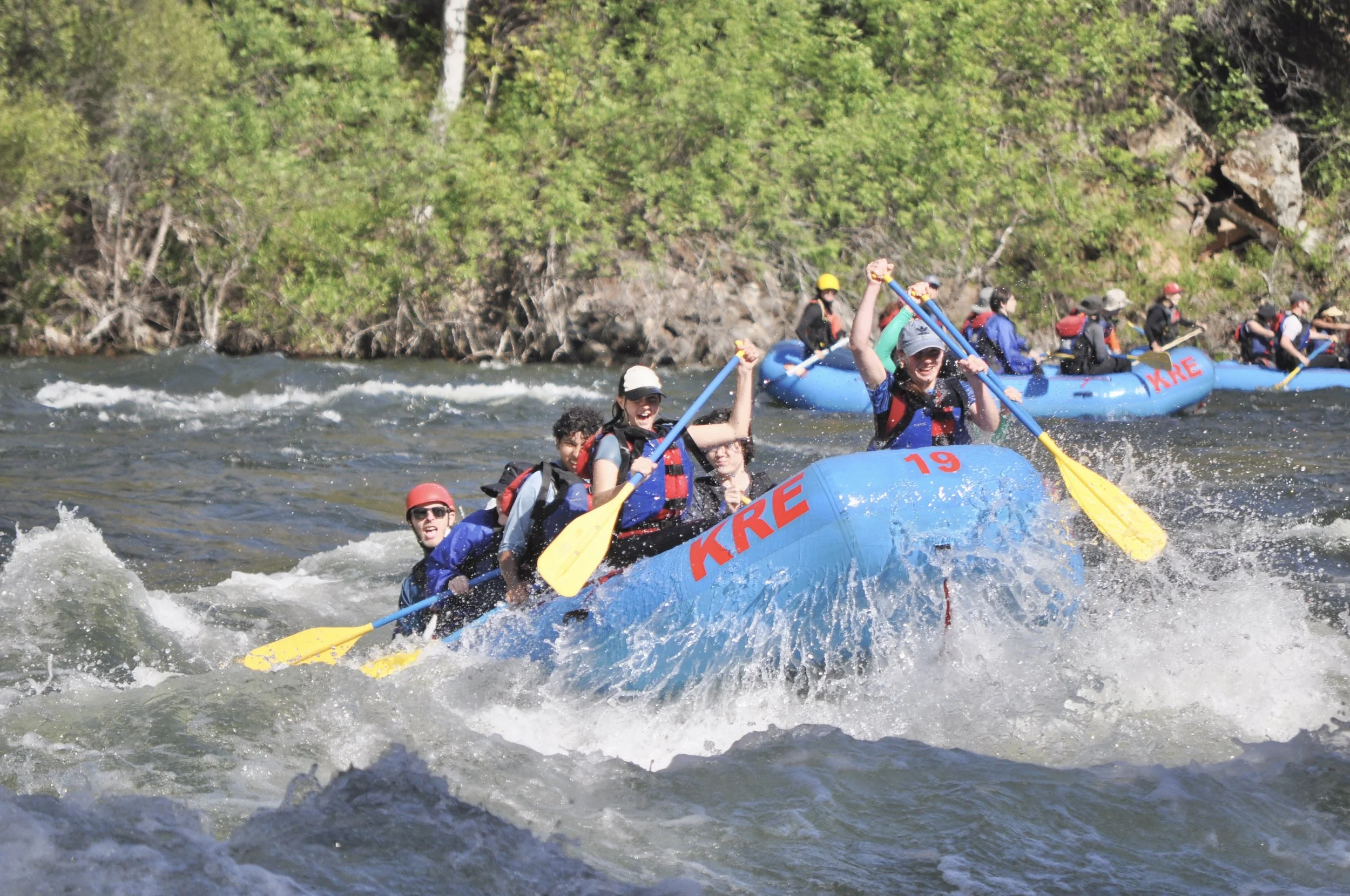 A group of people white water rafting on a river, wearing life jackets and helmets, with lush green trees and rocks in the background.