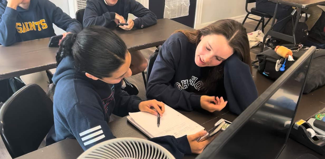 Two young women sitting at a table in a classroom, looking at a smartphone together. One is writing in a notebook, and both are engaged in conversation.
