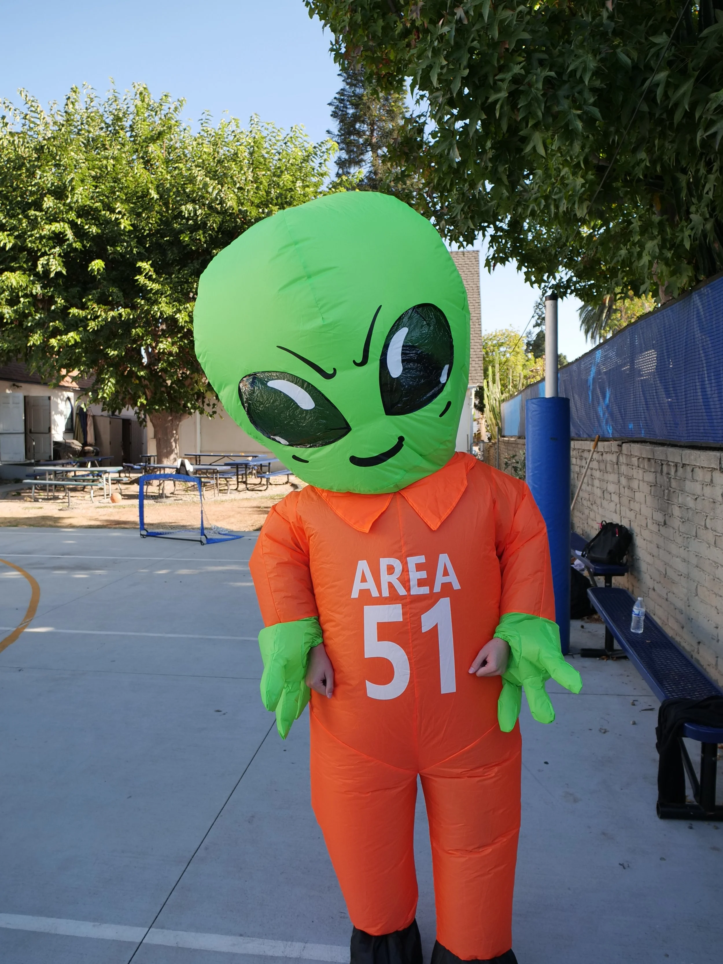 Person wearing an alien costume with a large green head and black eyes, orange jersey with the words 'AREA 51' on it, standing outdoors in a park or playground area.