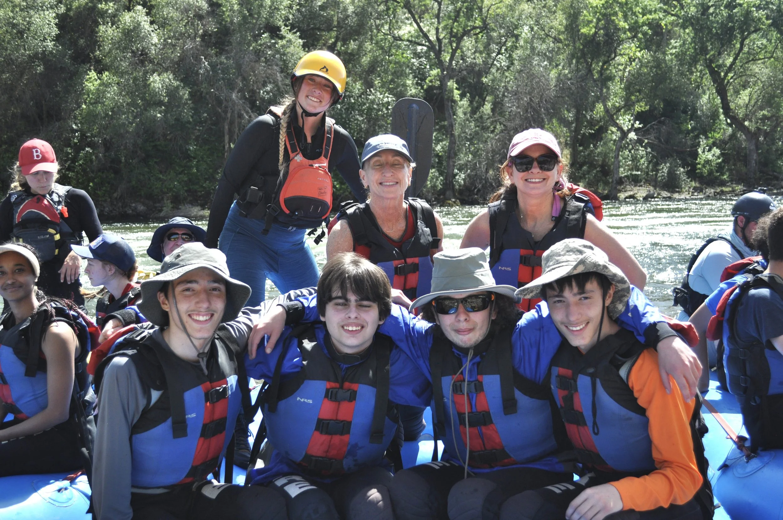 Group of smiling people in life jackets and helmets on a river, surrounded by trees.
