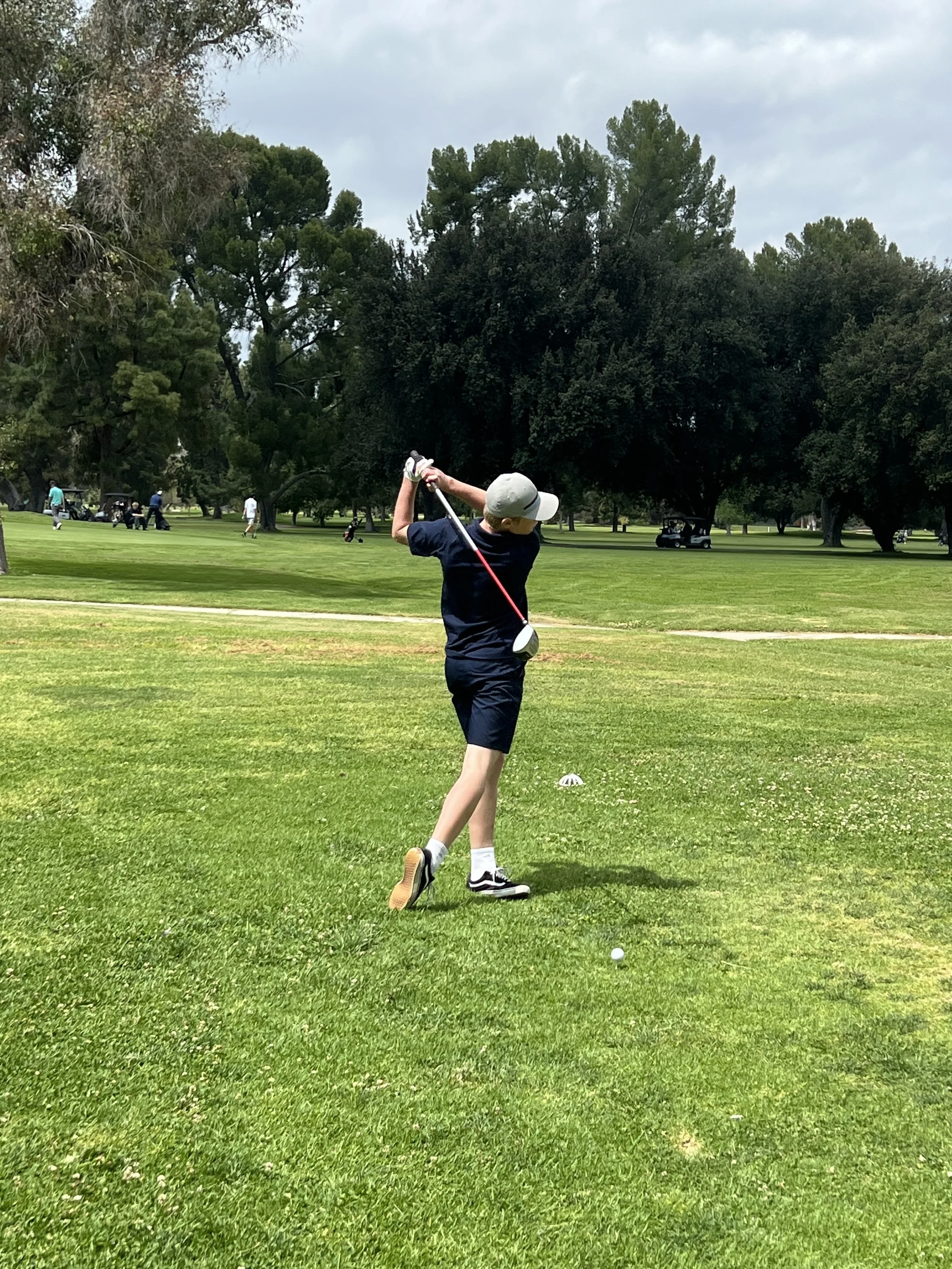 A boy swinging a golf club on a golf course with trees and golf carts in the background under a cloudy sky.
