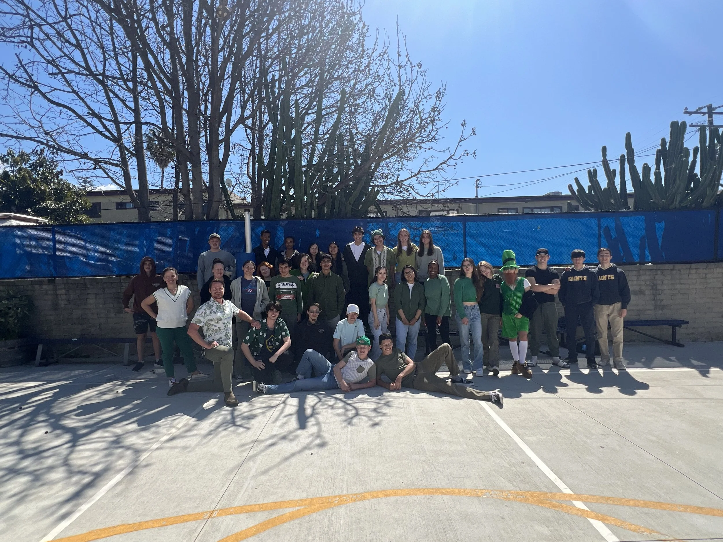 A large group of diverse young people standing and sitting outdoors on a sports court, with leafless trees, a blue fence, and some buildings in the background. The sky is clear and sunny.