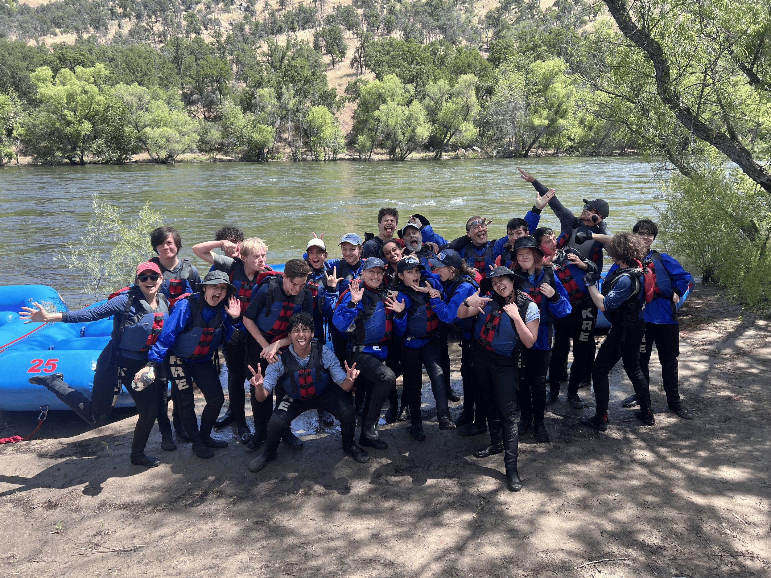Group of people, including children and adults, dressed in life jackets and outdoor gear, posing happily by a river with green trees and hilly landscape in the background.