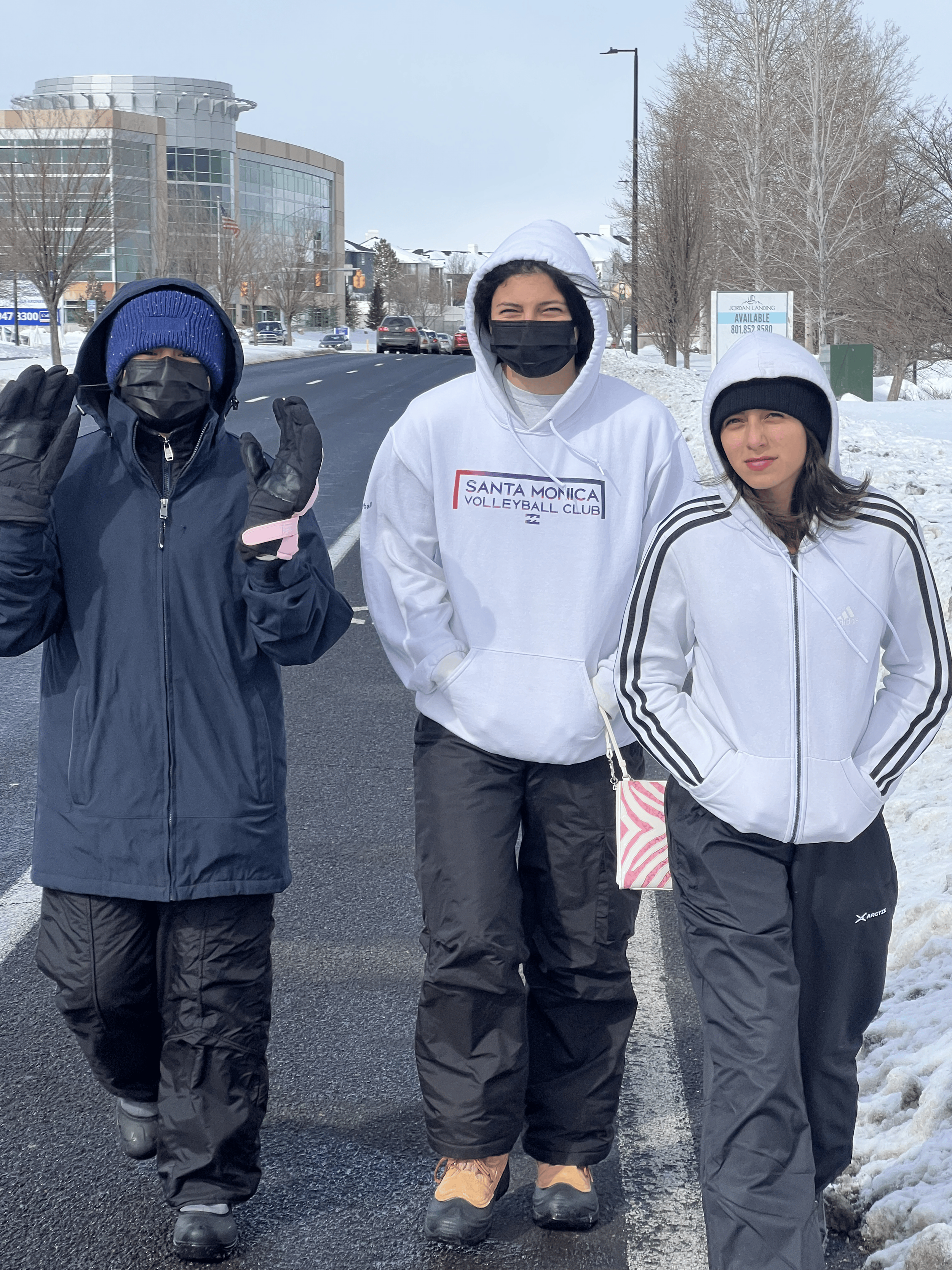 Three people walking outdoors in winter wearing jackets, hoodies, hats, and masks, with snow on the ground and a building in the background.