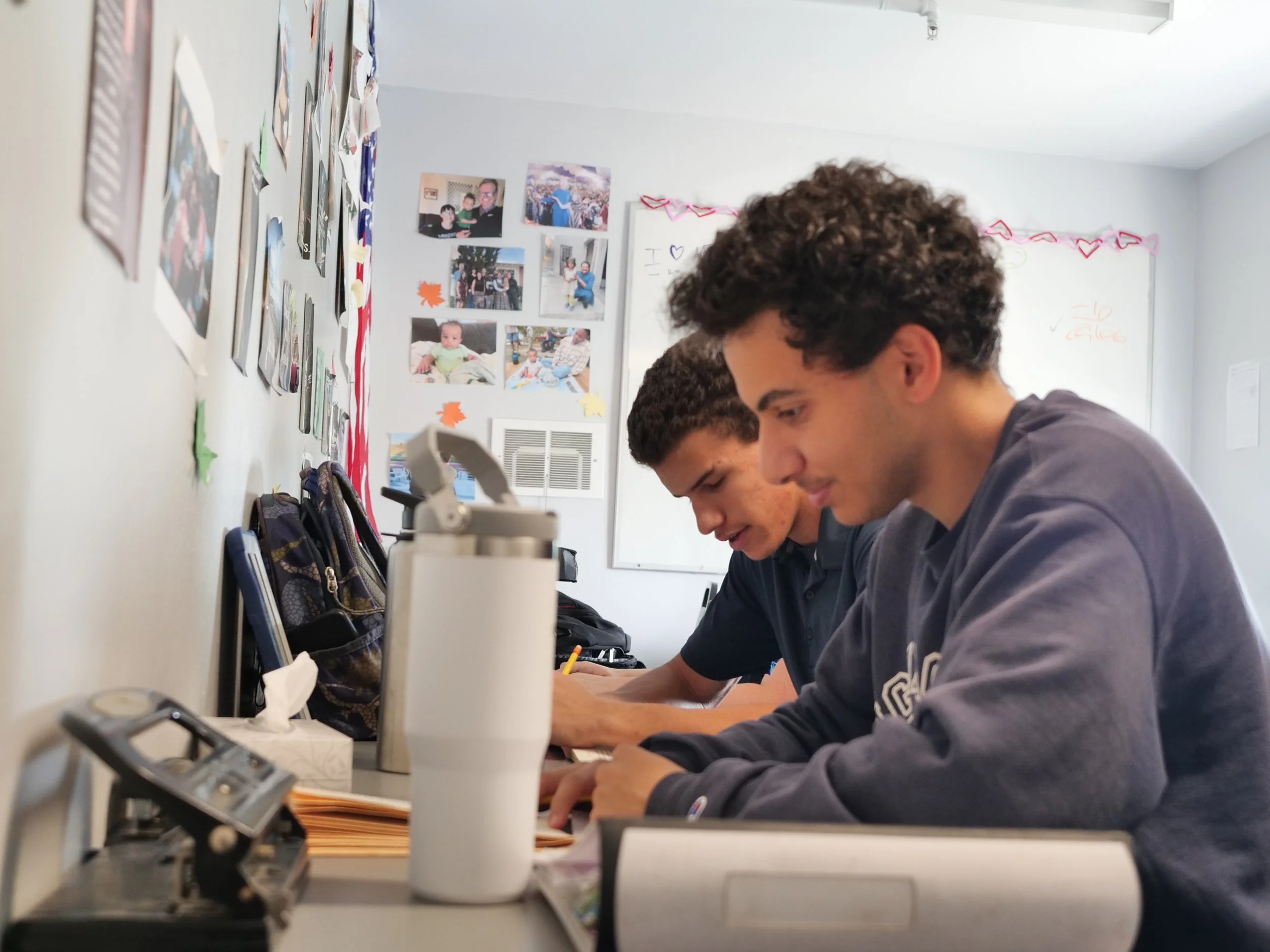 Two teenage boys sitting at a desk, studying with backpacks and school supplies ahead of them in a classroom decorated with photographs and posters.