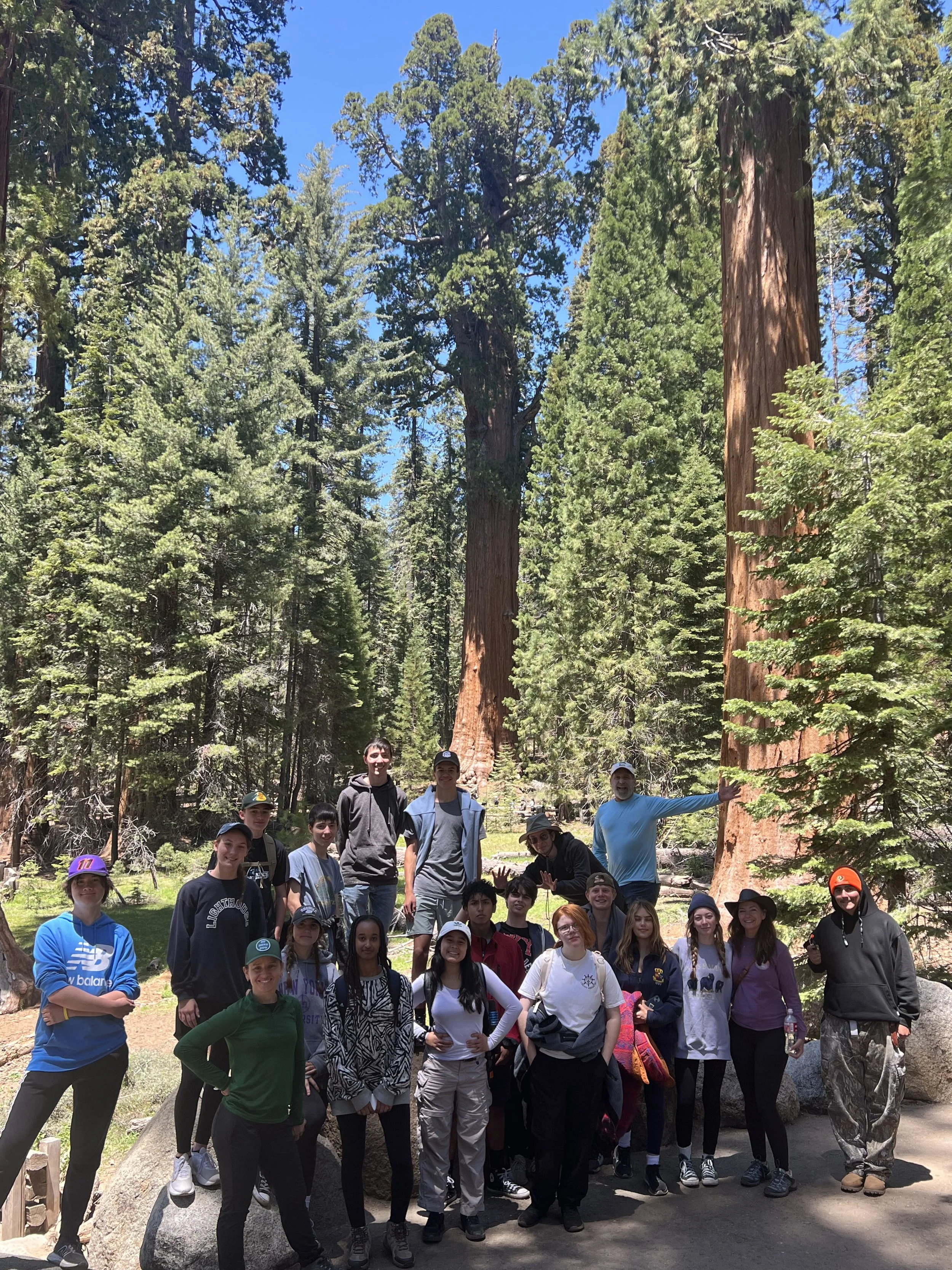 A group of people, including children and adults, posing together outdoors in a forested area with tall trees and blue sky visible. Some individuals are wearing hats and casual clothing, and they appear happy and excited.