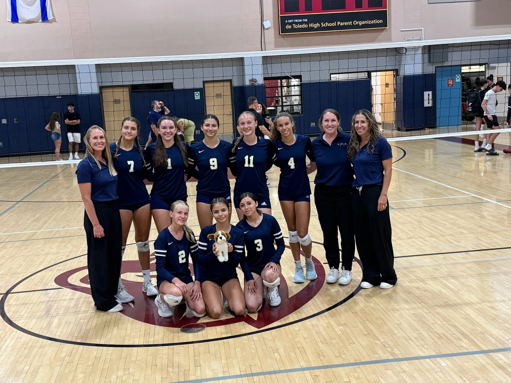 A group of seven young women in navy blue volleyball uniforms and two women in casual clothes standing in front of a volleyball net inside a gymnasium. Behind them, other people are visible, some standing and chatting, others preparing for volleyball play. The gym has a light-colored wood floor and a sign on the wall that mentions the Toledo High School Parent Organization.