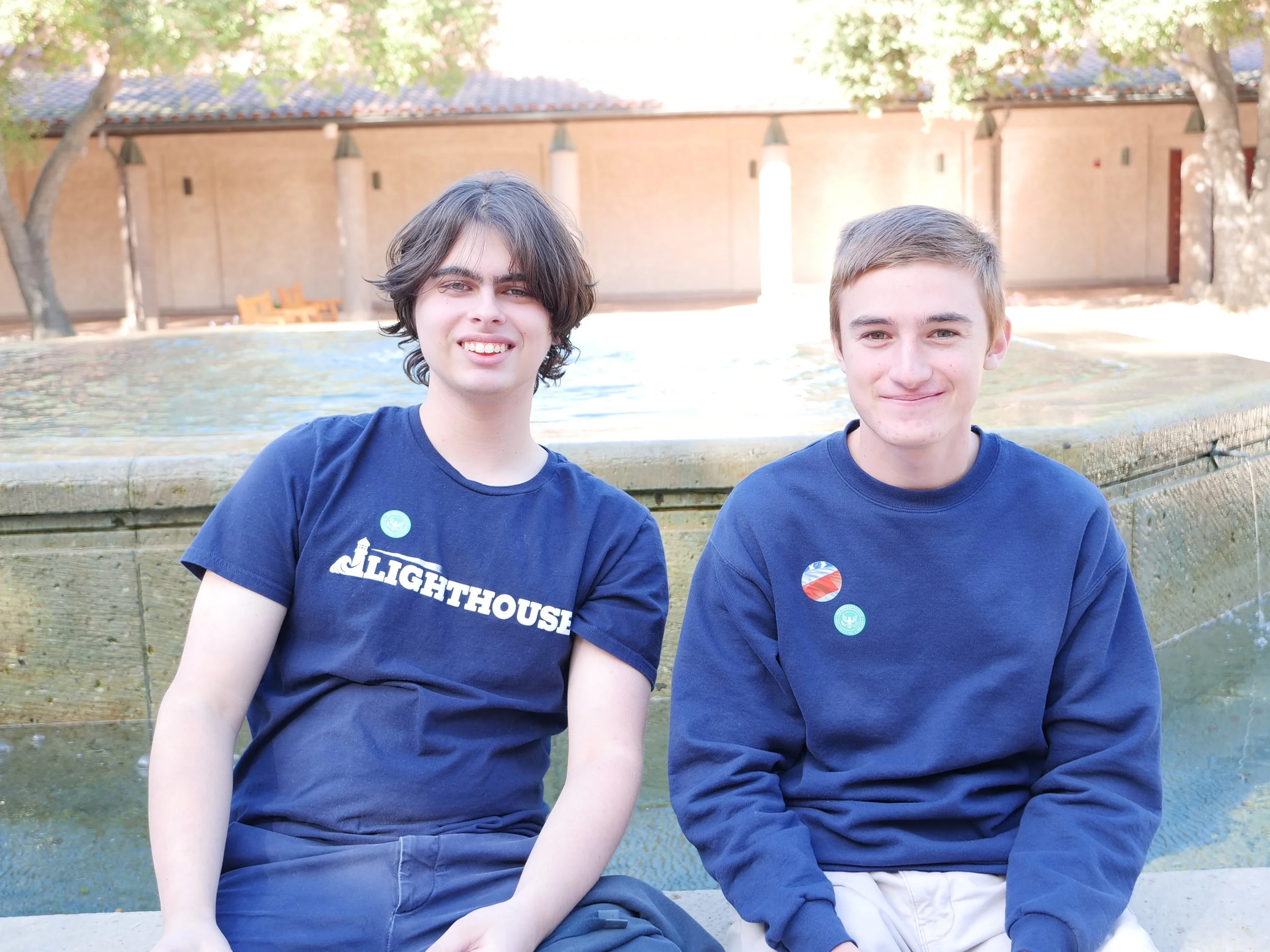 Two young men sitting outdoors near a fountain with trees and a brick building in the background.