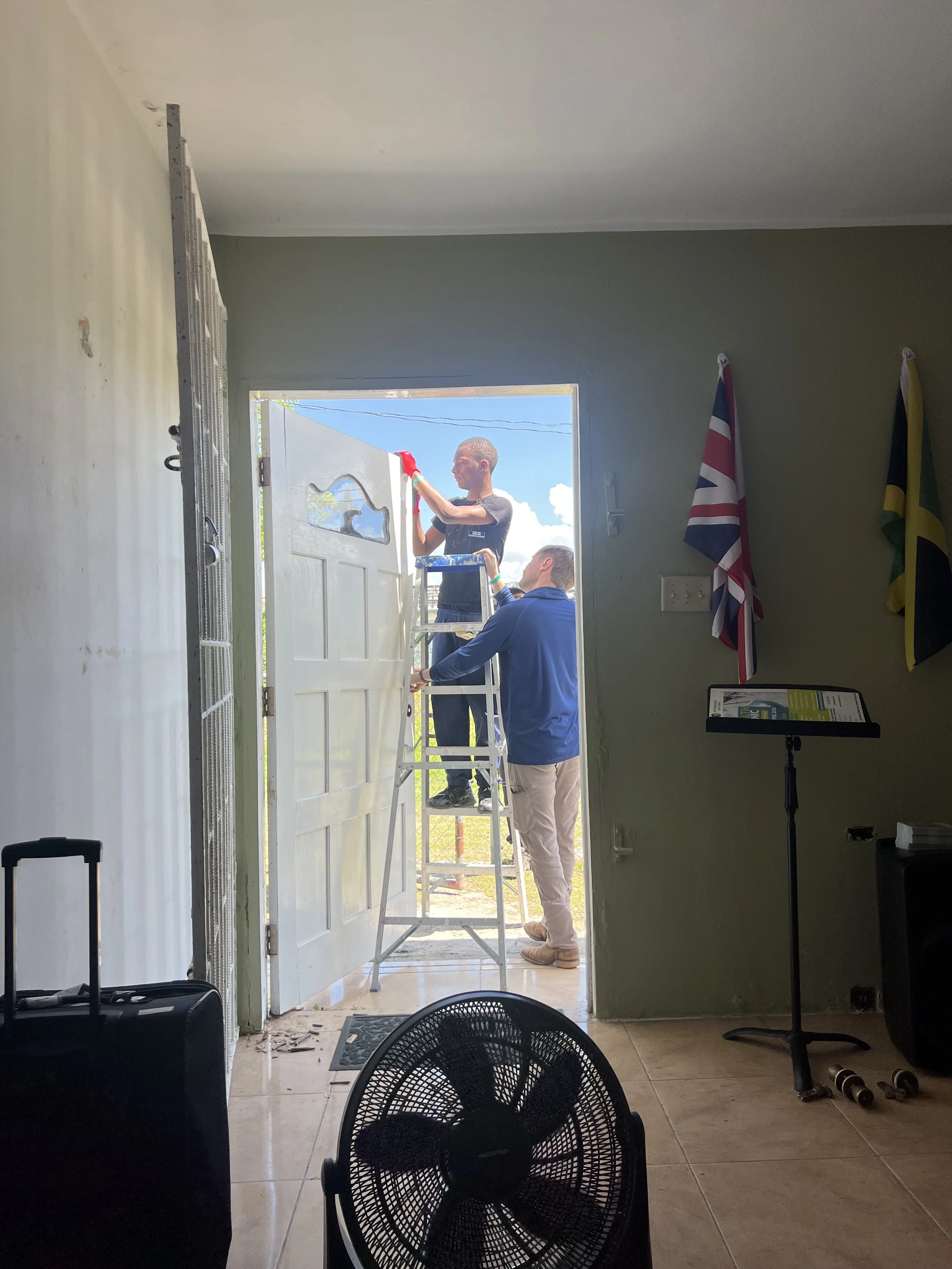 People installing a white door using a ladder. Two men are working outside the house, one standing on the ladder and the other holding the ladder. Flags are hanging on the wall inside, and gym weights are on the floor.