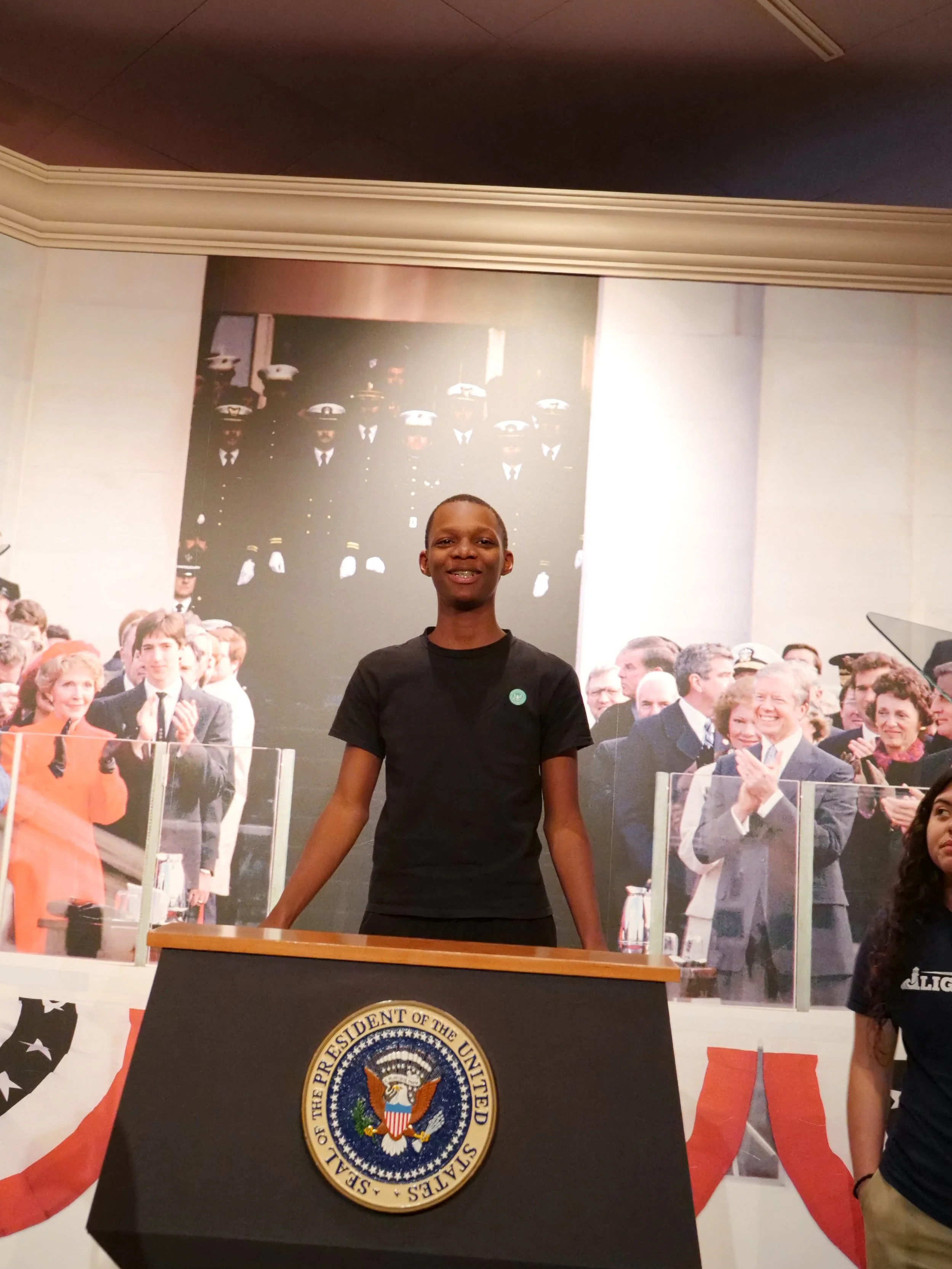 A young man standing behind a presidential podium in front of a large photograph of a formal event, with people clapping and smiling in the background.