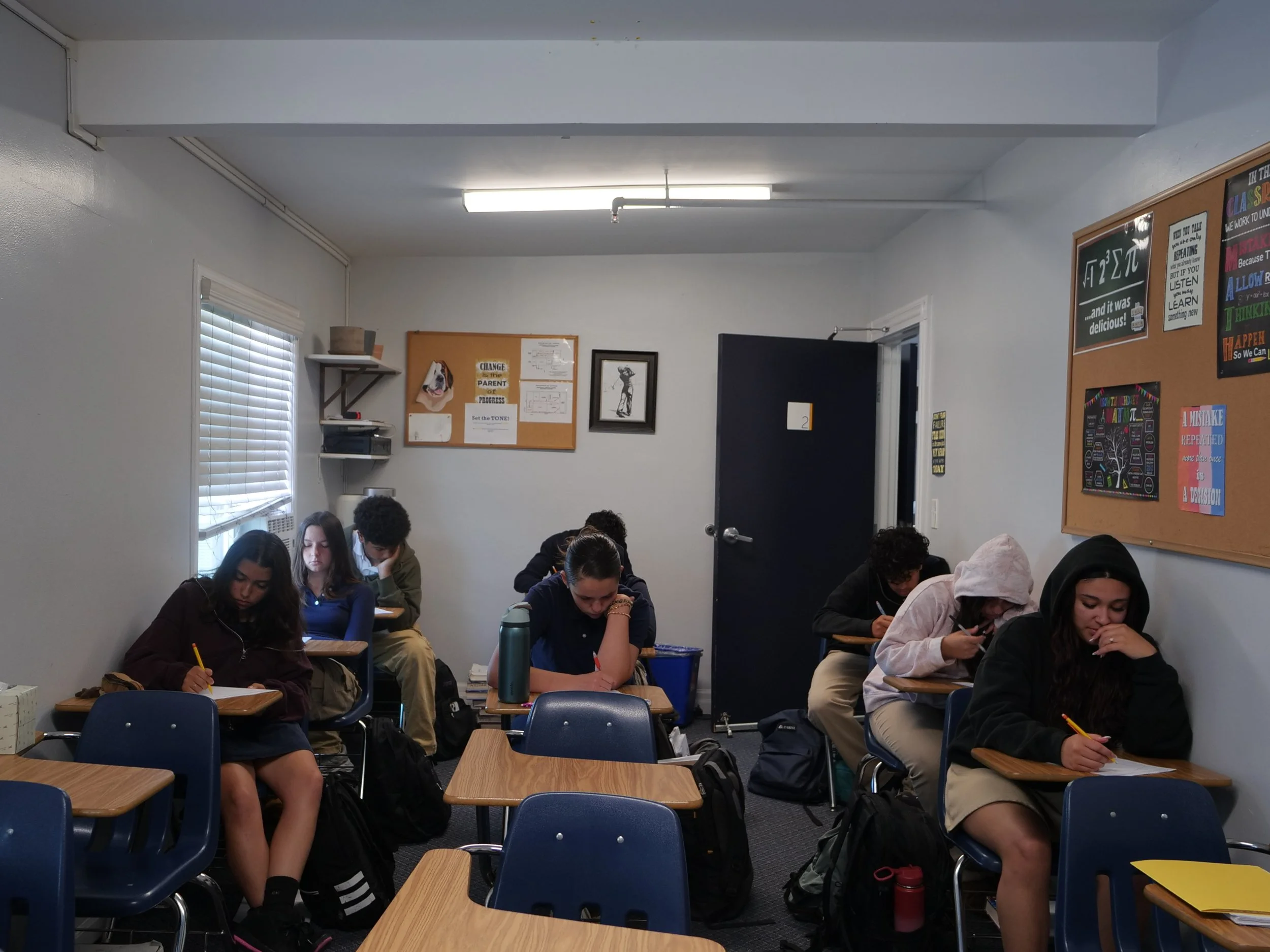 A classroom scene with eight students seated at desks, focused on their work, with white blinds on the window to the left and motivational posters on the wall to the right. The students are wearing school uniforms or casual clothing.