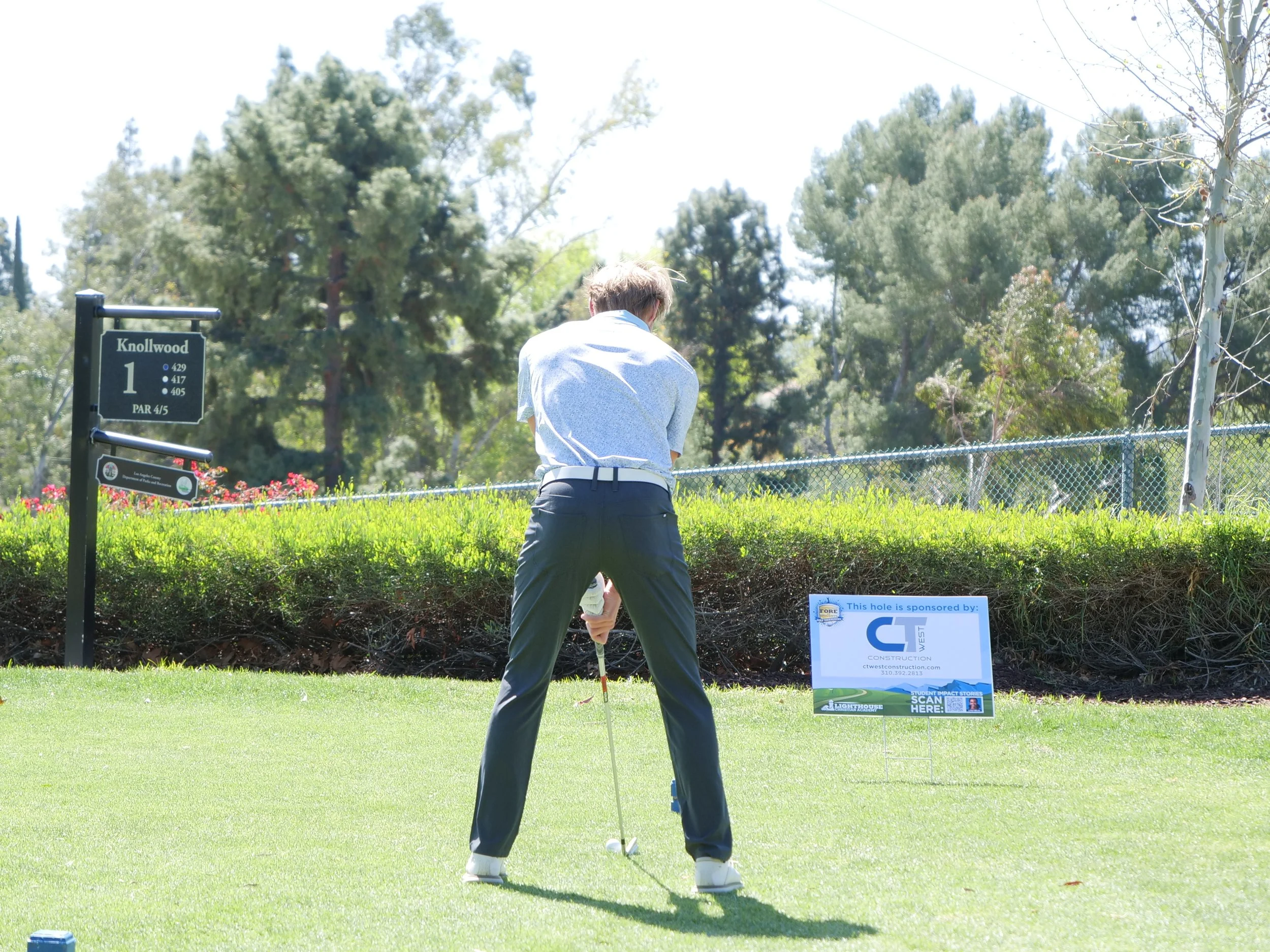 A man playing golf on a green course, preparing to putt, with trees and signage in the background.