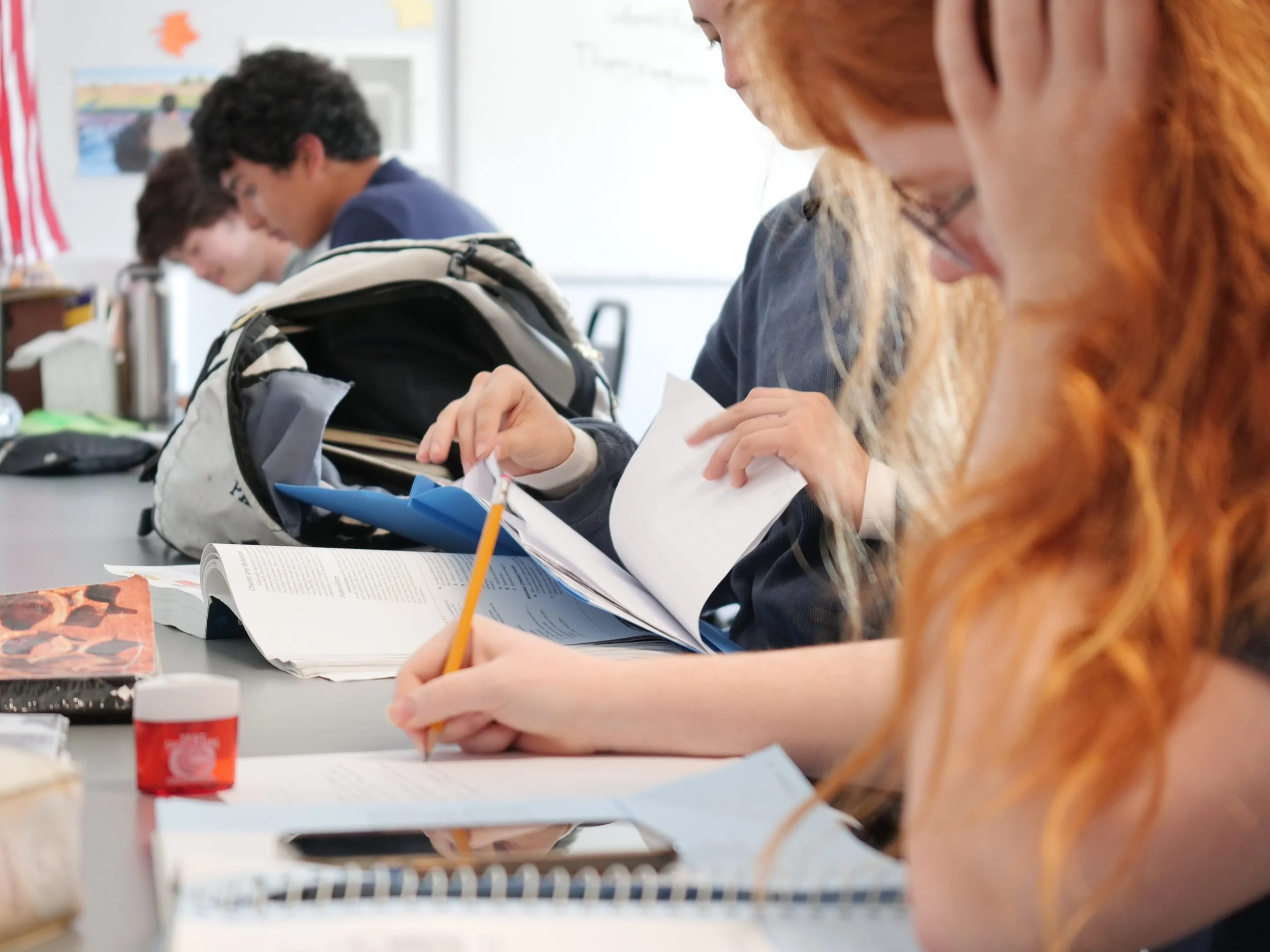 Students in a classroom working on assignments, with school supplies and backpacks on the desk.