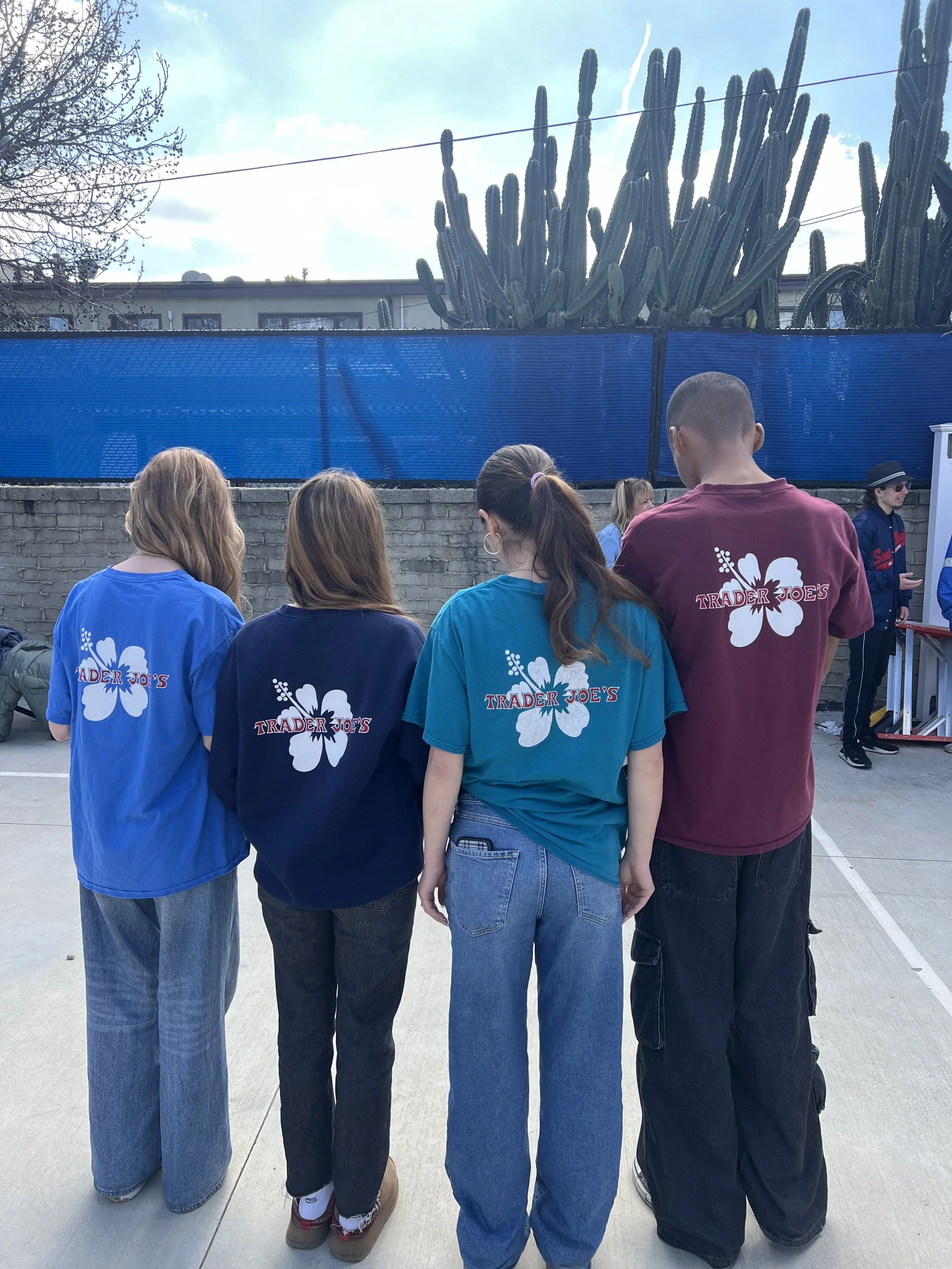 A group of people standing outdoors with backs to the camera, wearing blue and maroon shirts with a white hibiscus flower and the words "Trader Joe's" on the back. They are in front of a blue fence and a large cactus, with a clear sky overhead.