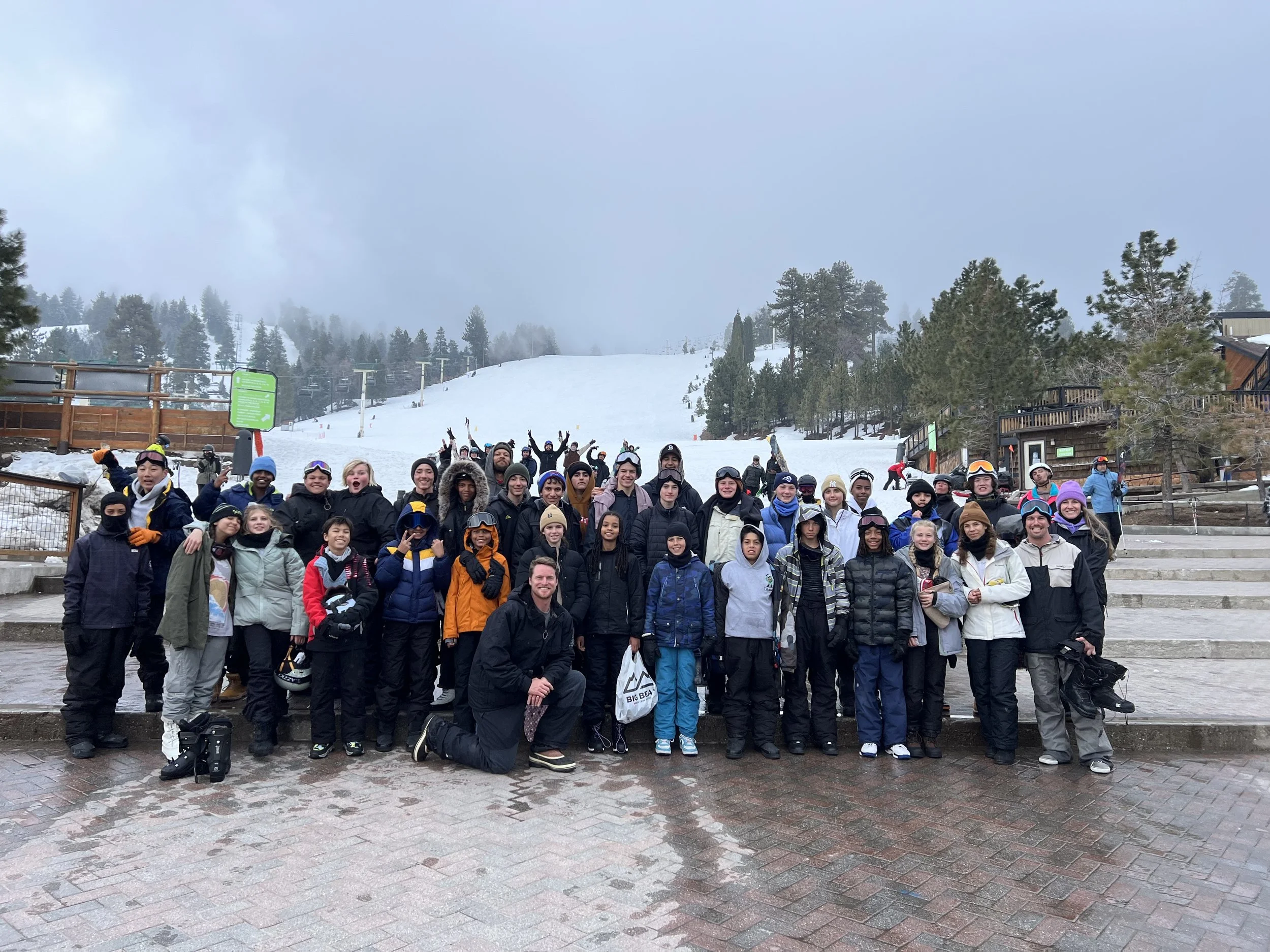Group of people in winter clothing posing on a step at a snowy ski resort, with snowy slopes and trees in the background.