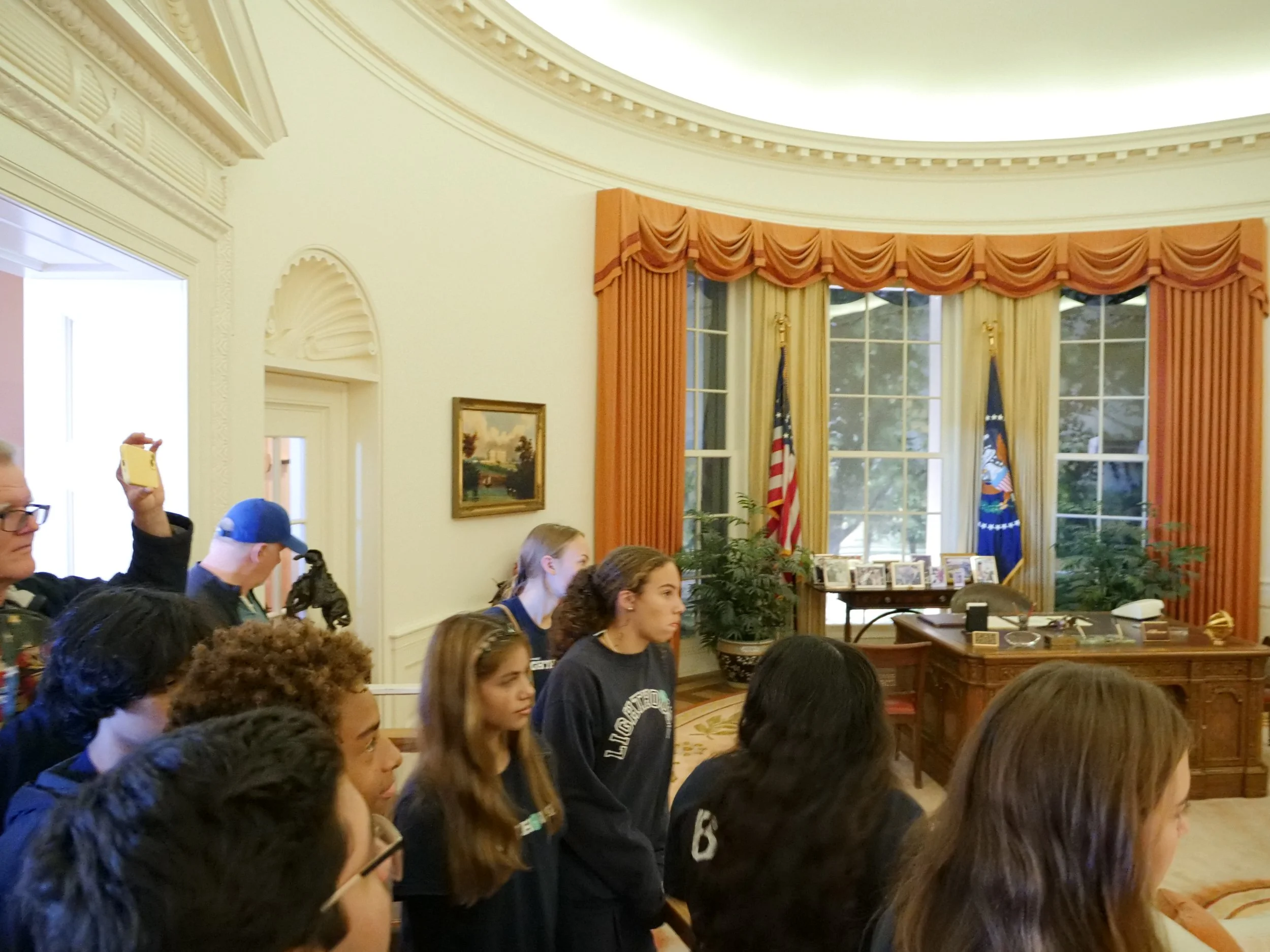 A group of young people, mostly women, are standing close together in a room with ornate curtains, flags, and framed photographs on a table. They are facing forward and appear to be listening attentively to an event or presentation.