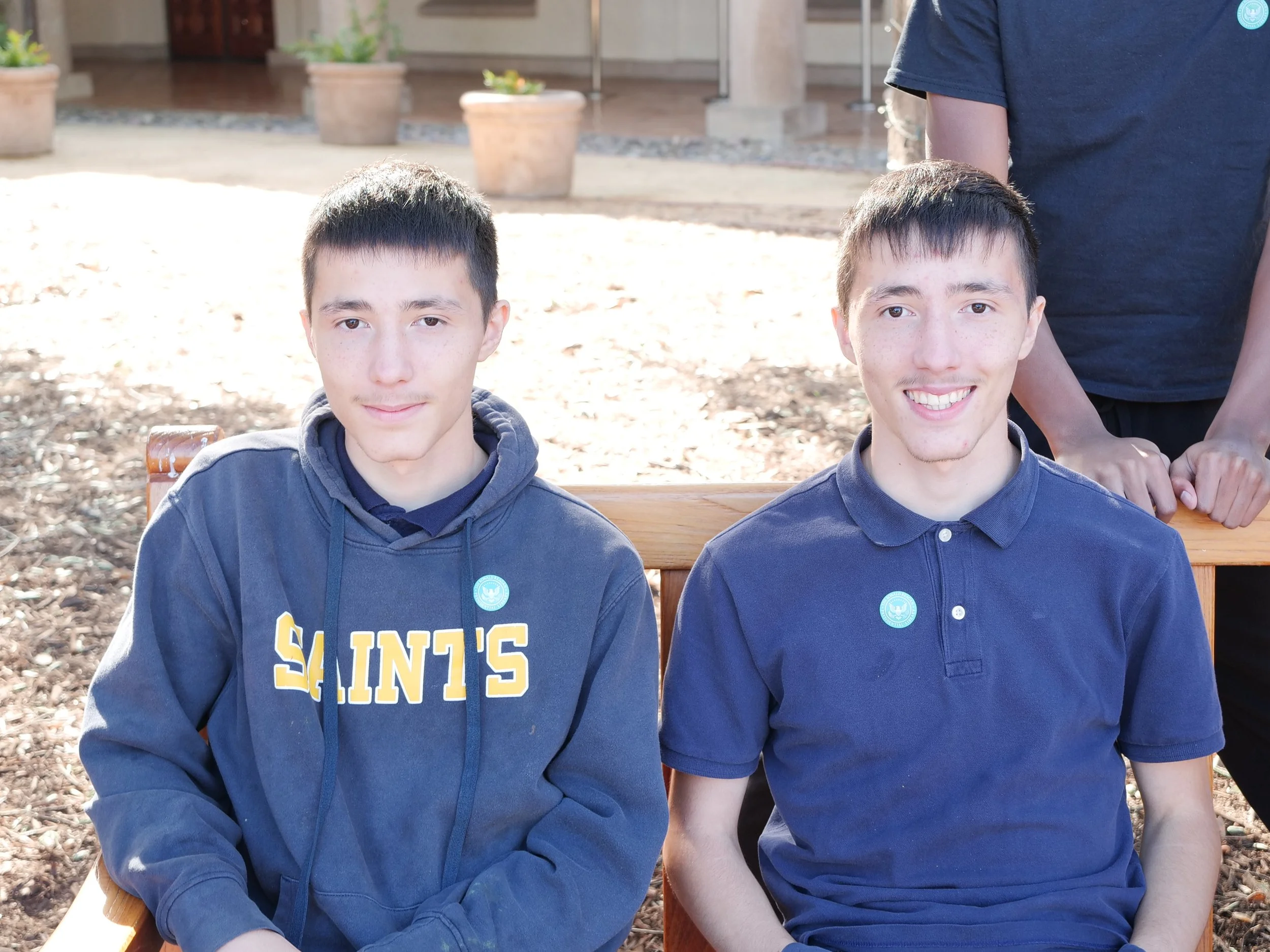 Two young men sitting on a bench outdoors, wearing navy blue shirts, with a blurred background of potted plants and a building.