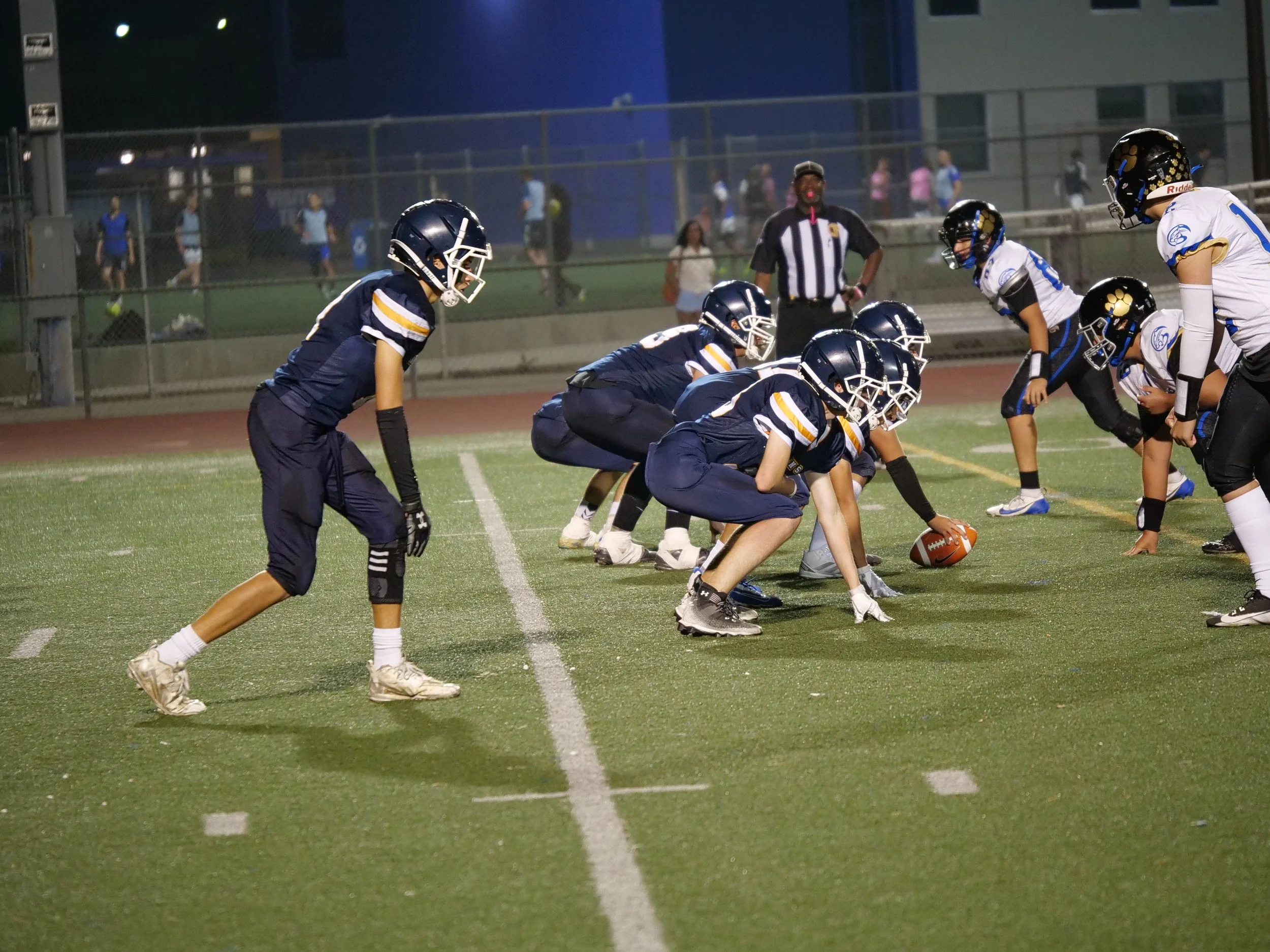 American football players in a ready stance on the field during a game at night, with officials and spectators visible in the background.