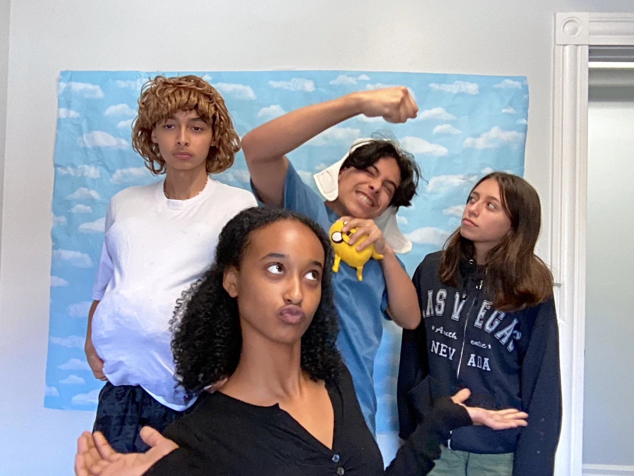 Group of five young people posing in front of a cloud-patterned backdrop, with one person flexing arm muscle, one making a pouty face, and others looking in various directions.