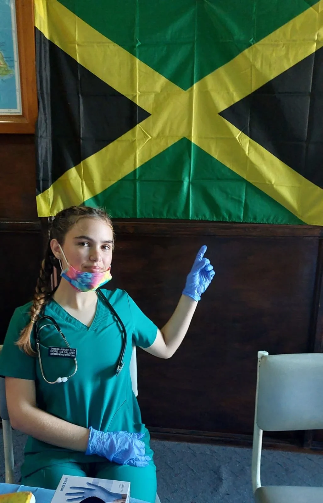 A young woman dressed as a nurse is sitting at a table, wearing scrubs, a stethoscope, blue gloves, and a colorful face mask. She is pointing upward with her right index finger. Behind her is a Jamaican flag hanging on a dark wooden wall.