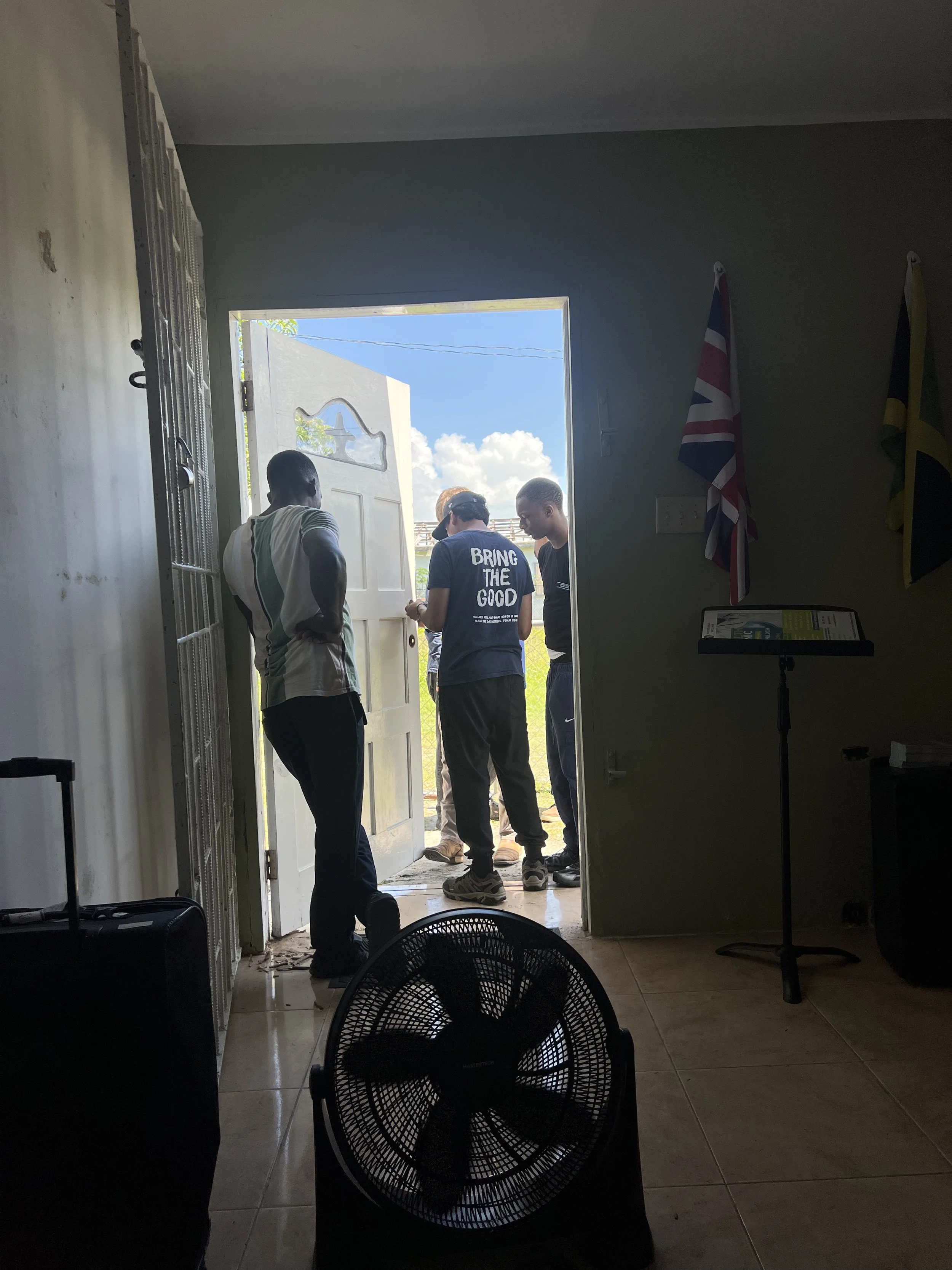 Three people standing outside a doorway, two men and one woman, with one person wearing a shirt that says 'Bring the Good,' as sunlight streams in from the open entrance.